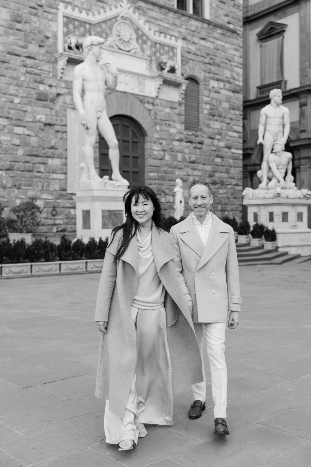 A couple in light colored coats walking together in front of historic stone architecture with classical statues.