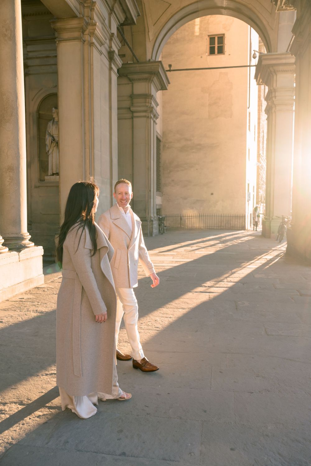 Sunlit archways in a historic stone building cast dramatic shadows on the ground while someone walks through.