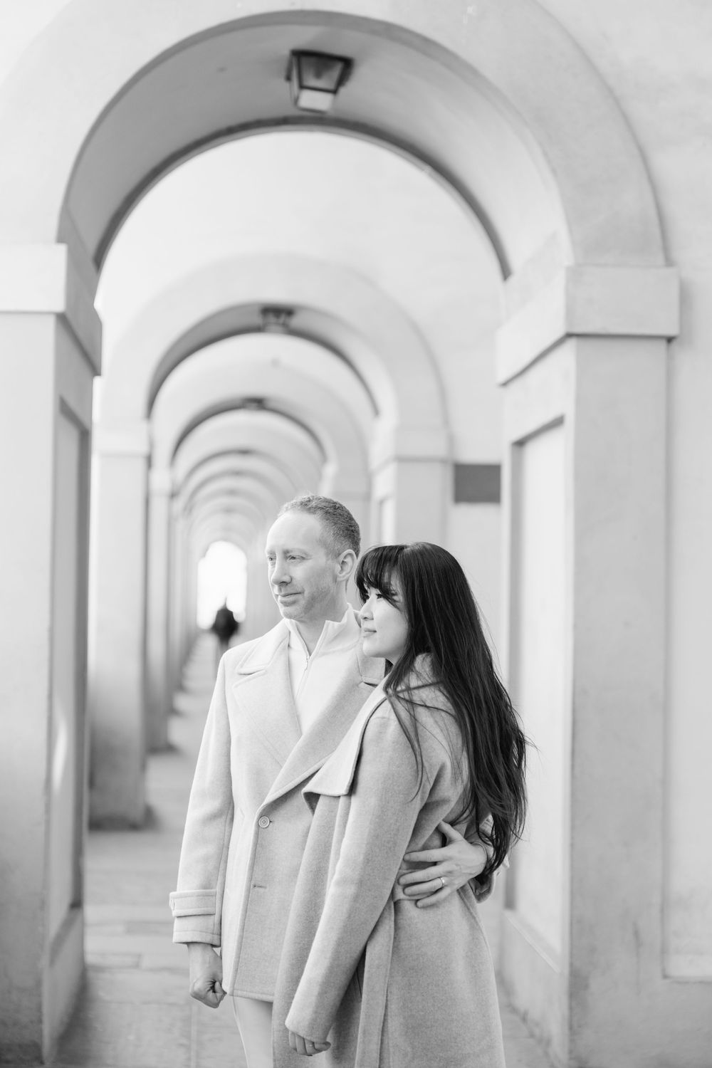 A couple in light coats embraces while standing in an elegant arched colonnade hallway in black and white.