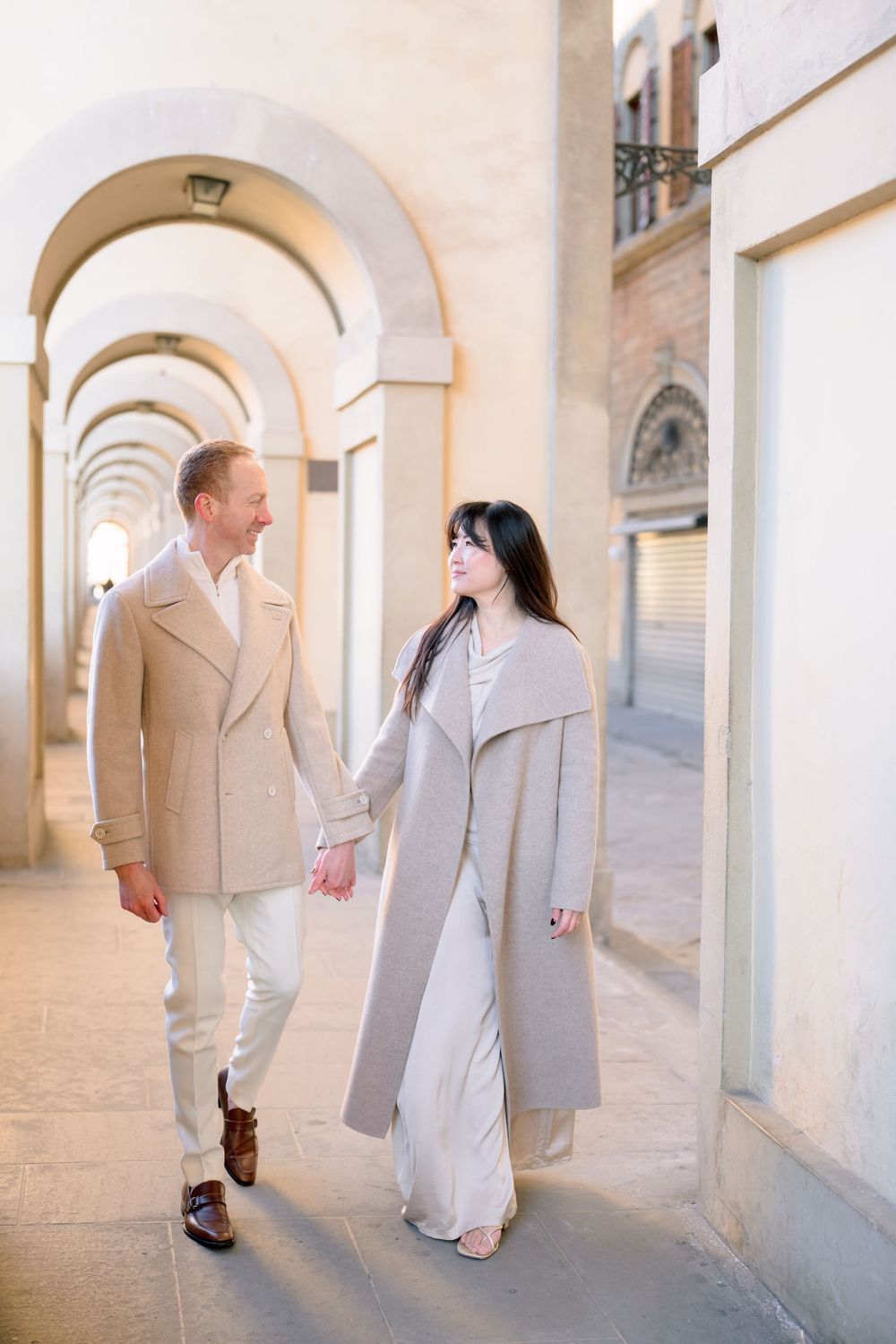 A couple in light colored formal attire walks through an elegant arched corridor with stone walls and columns.
