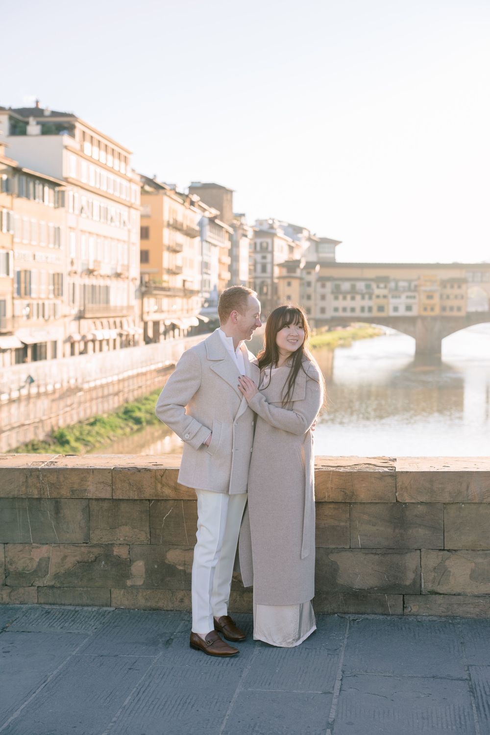 Couple in matching beige outfits poses along stone wall overlooking Florence's Arno River and Ponte Vecchio bridge.