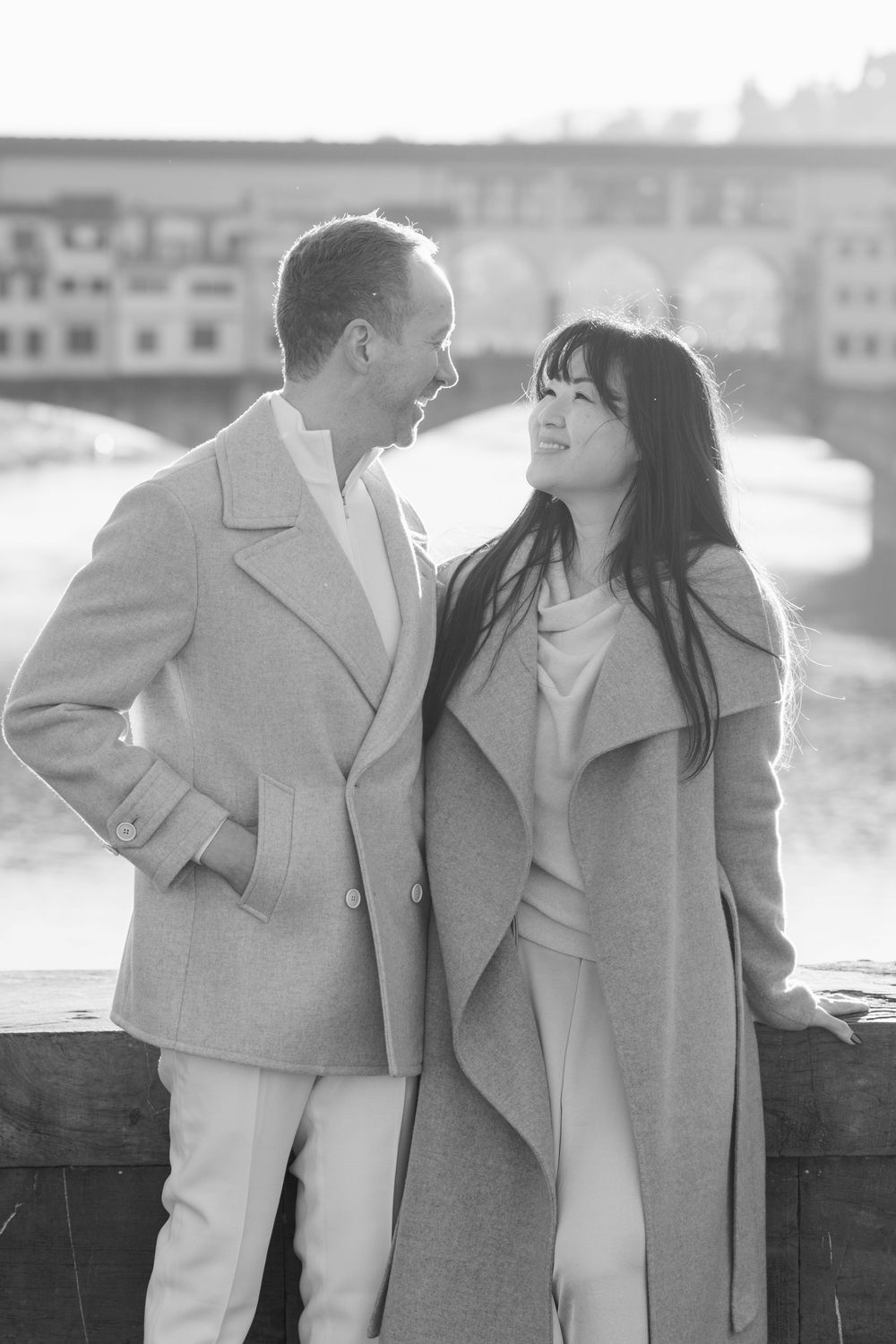 A romantic black and white photo series showing a couple in matching light coats embracing by a waterfront.