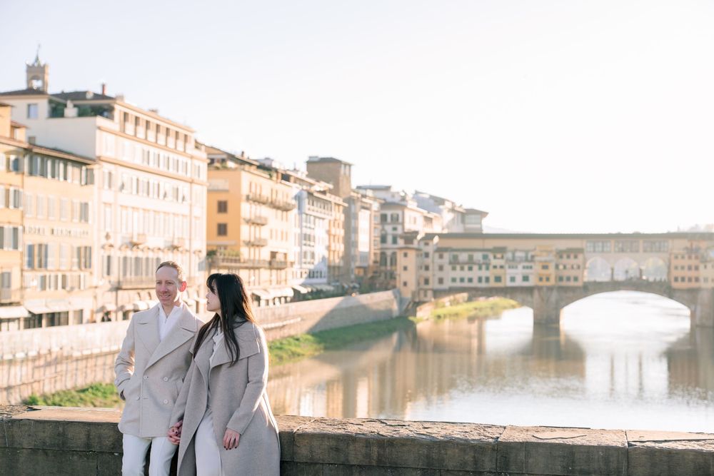 Romantic couple embraces while sightseeing along the Arno River in Florence, Italy with historic buildings in background.