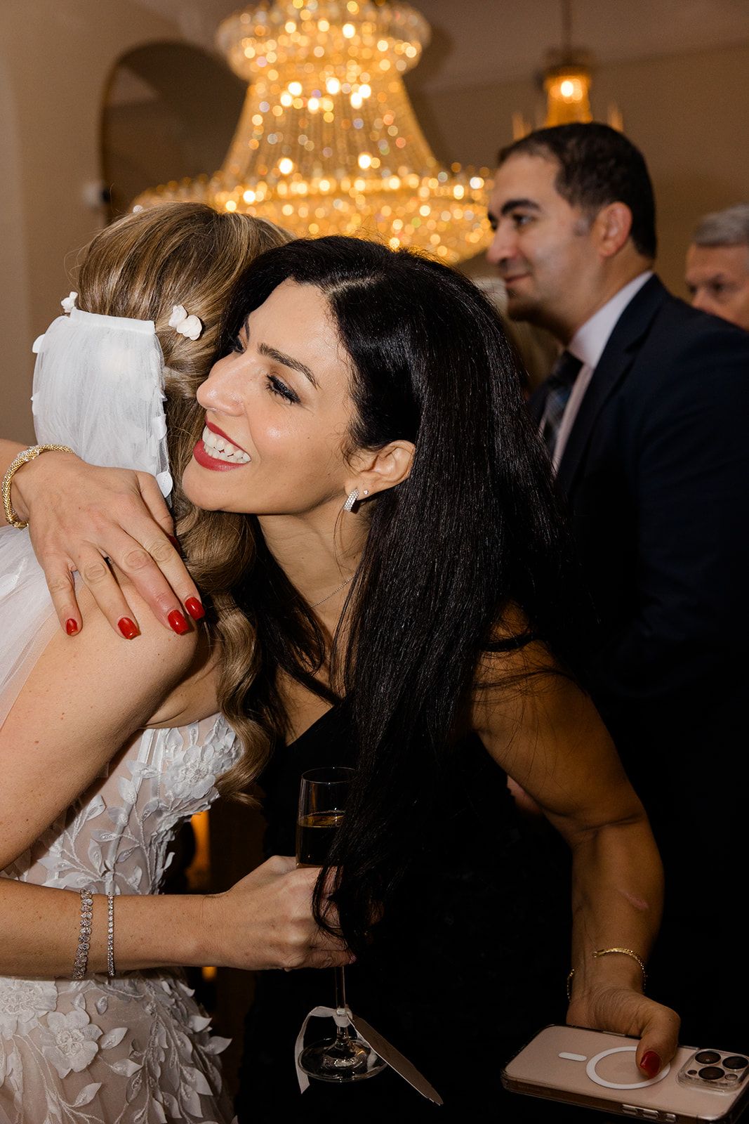 Two women sharing a joyful moment at an elegant event with chandeliers glowing in the background.