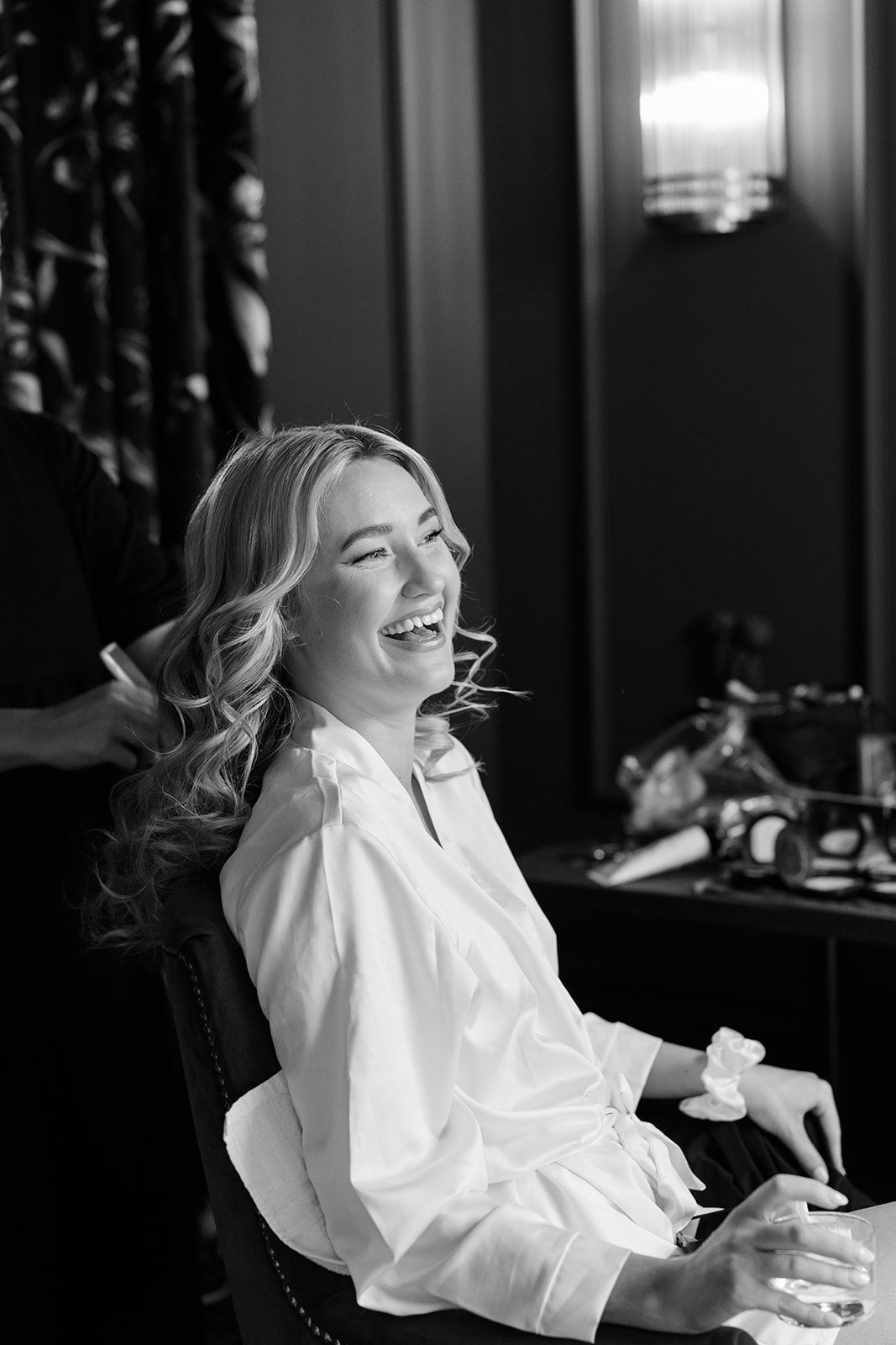 A person in a white shirt laughs joyfully while seated in a dark room, captured in black and white photography.