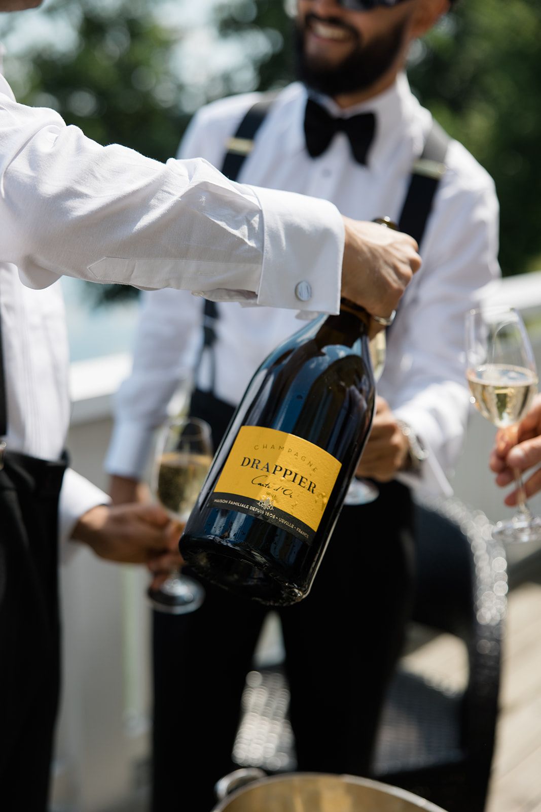 Servers in formal attire pour champagne from bottles at an elegant outdoor event.