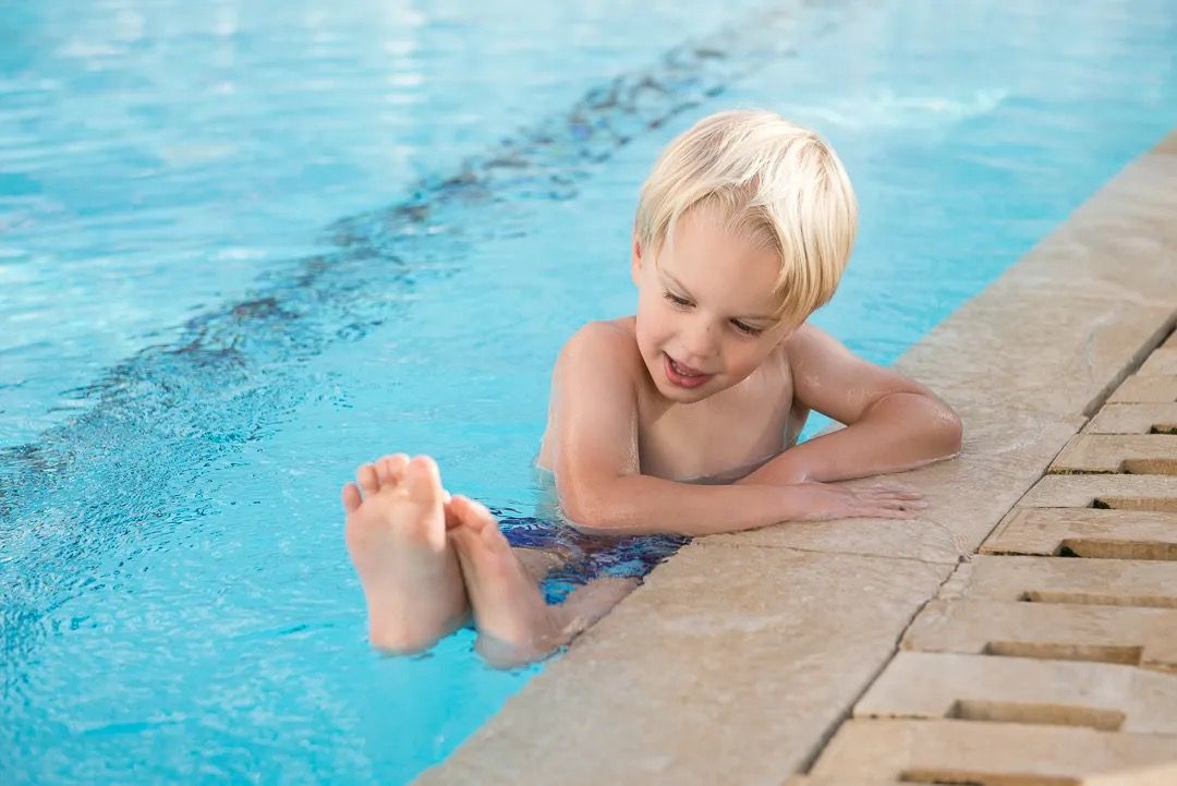 A young child sitting on the pool edge with feet in the water on a sunny day.
