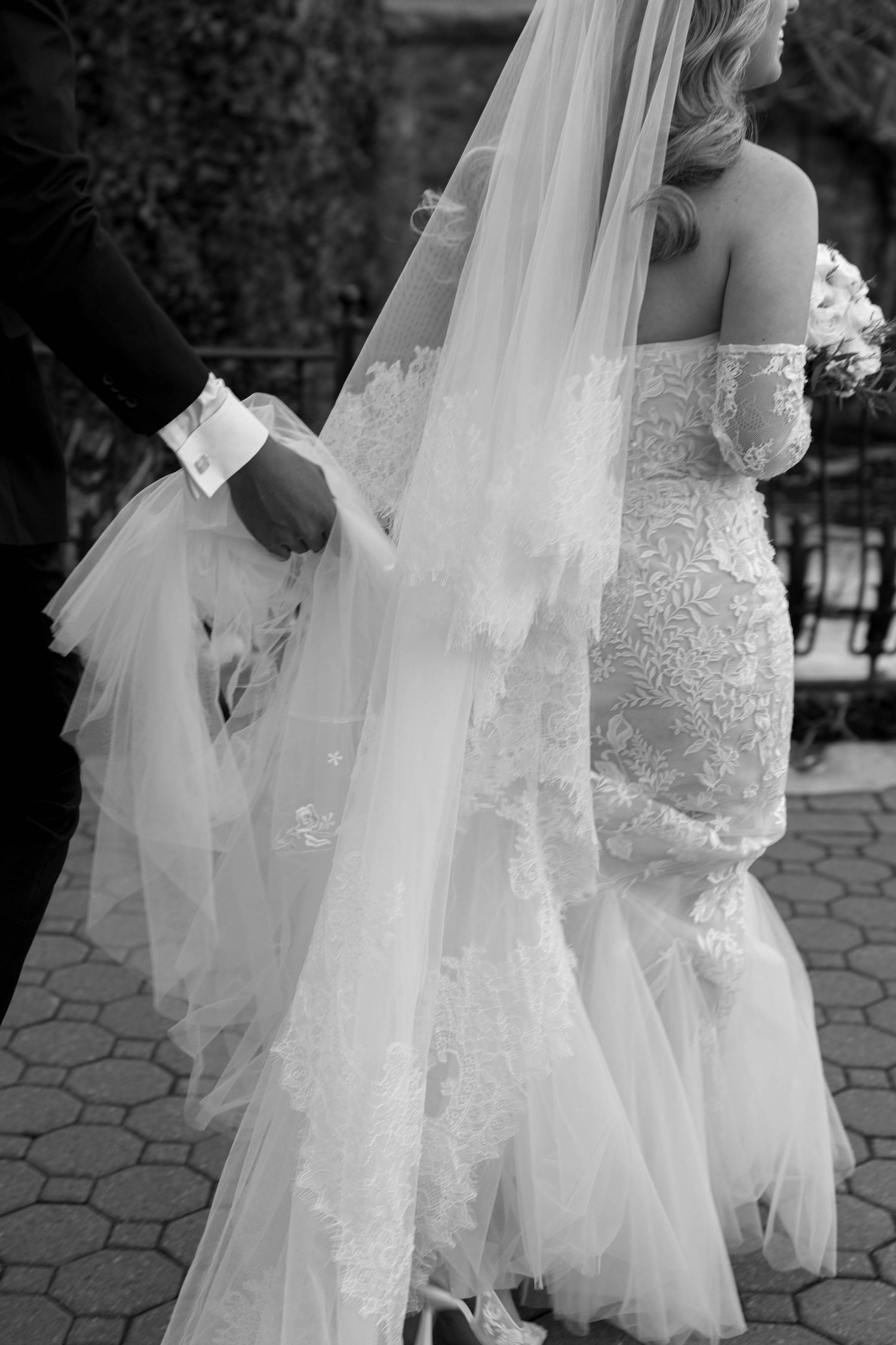 Romantic wedding couple poses outdoors with a white parasol and bouquet in a garden setting.