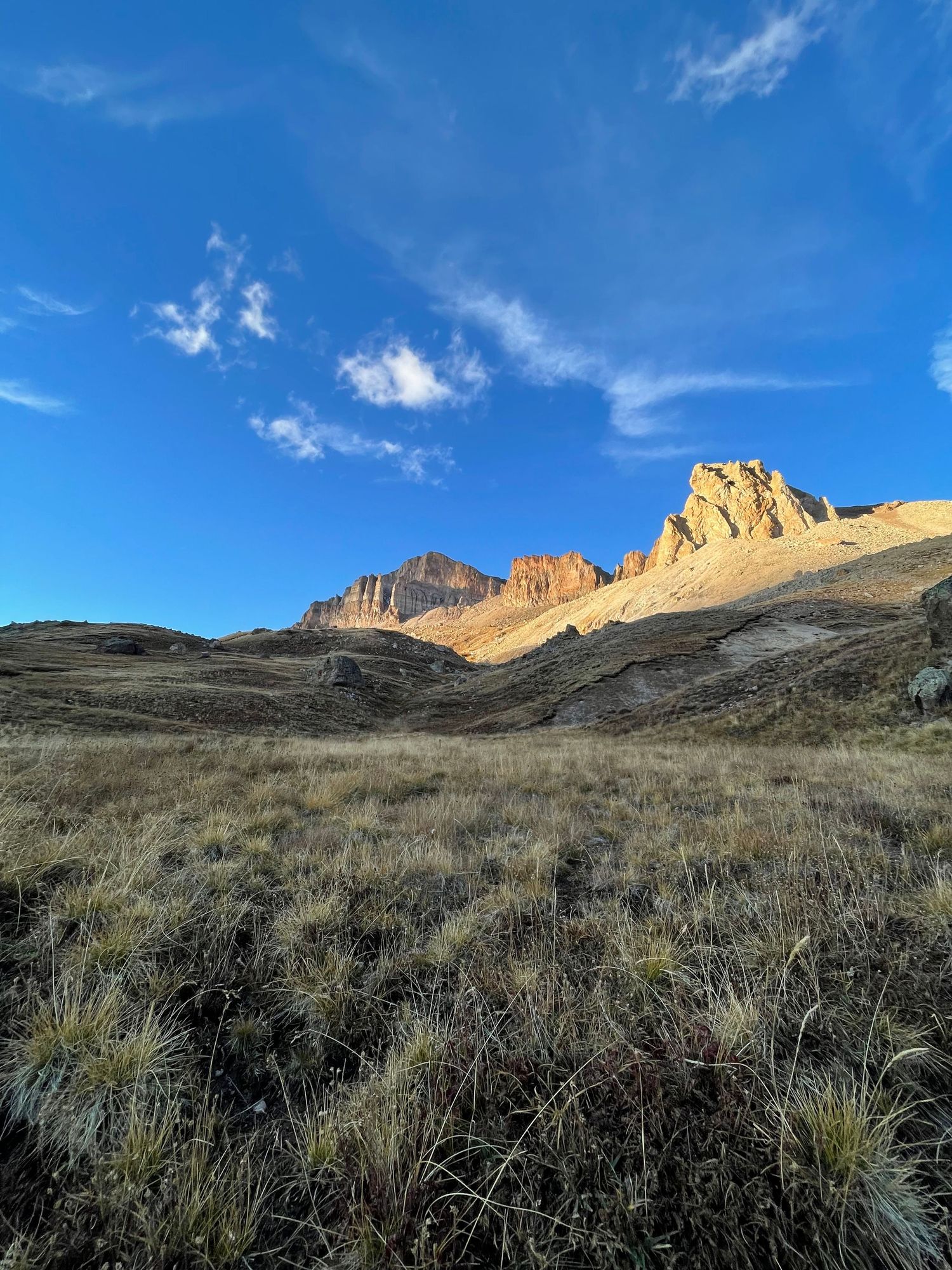 A panoramic mountain landscape with rugged peaks and scrubland under a bright blue sky with wispy clouds.