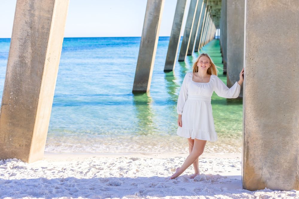 Family poses for beach photos under a pier on a sunny day with turquoise ocean water in the background.