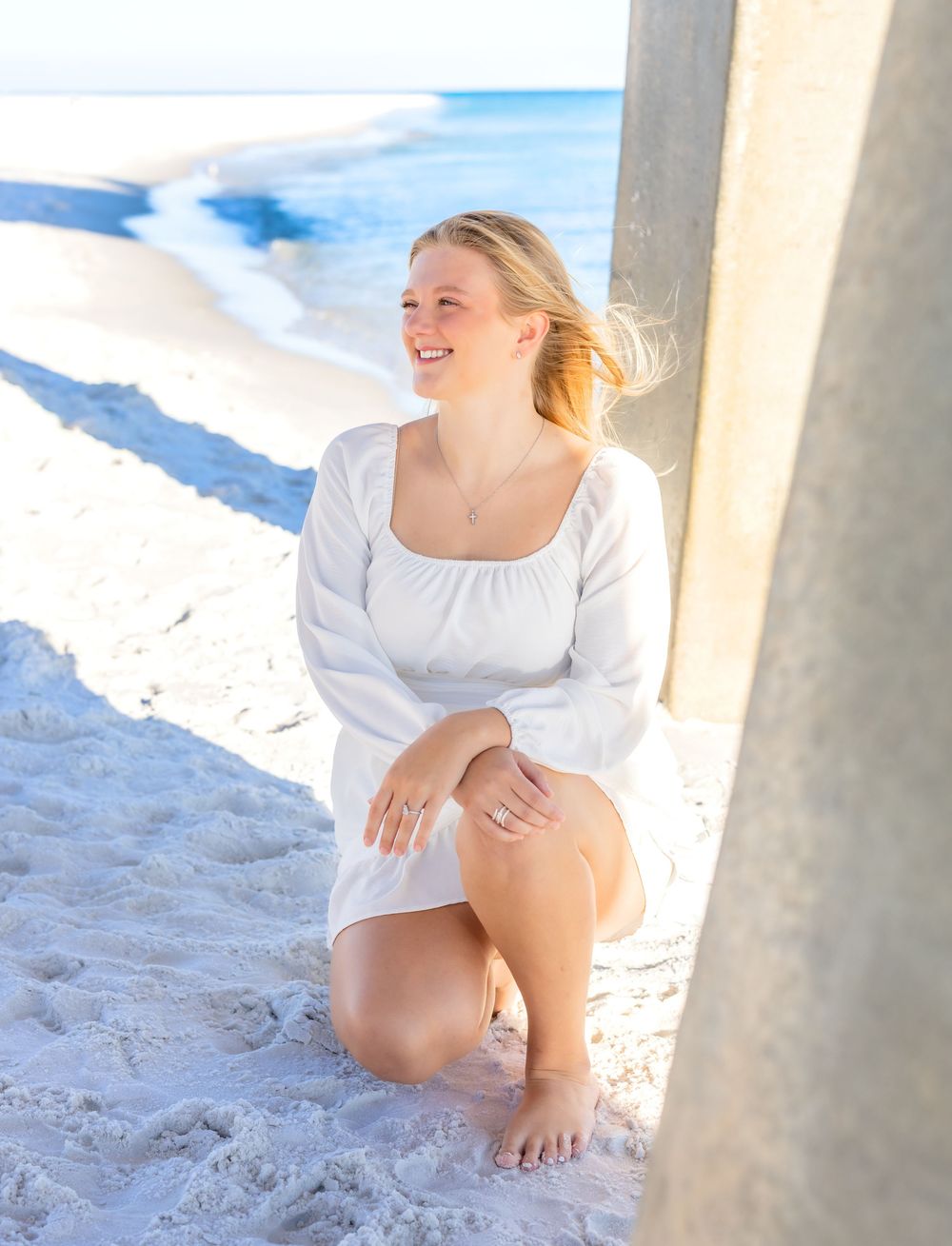 A person in a white dress sits gracefully on sandy ground near white pillars with snow-capped mountains in the background.