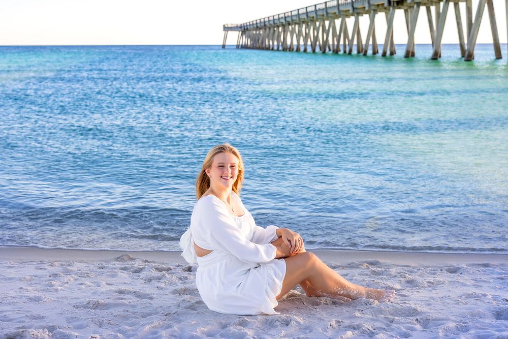 A person in a white dress poses on the beach with a pier in the background at sunset.