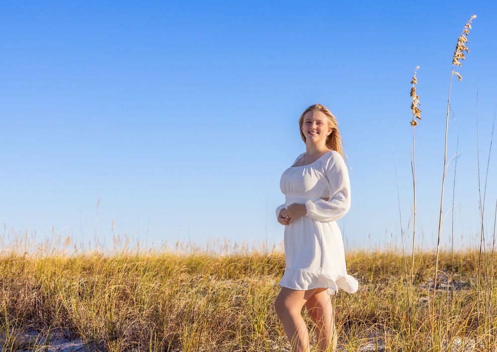 A person in a white dress stands in a golden field of tall grass against a bright blue sky on a sunny day.