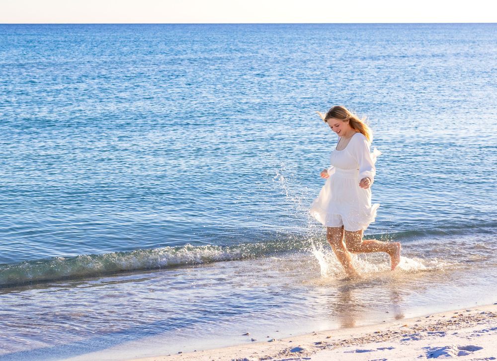 A person in a white dress walks along a scenic beach shoreline with calm blue waters and gentle waves.