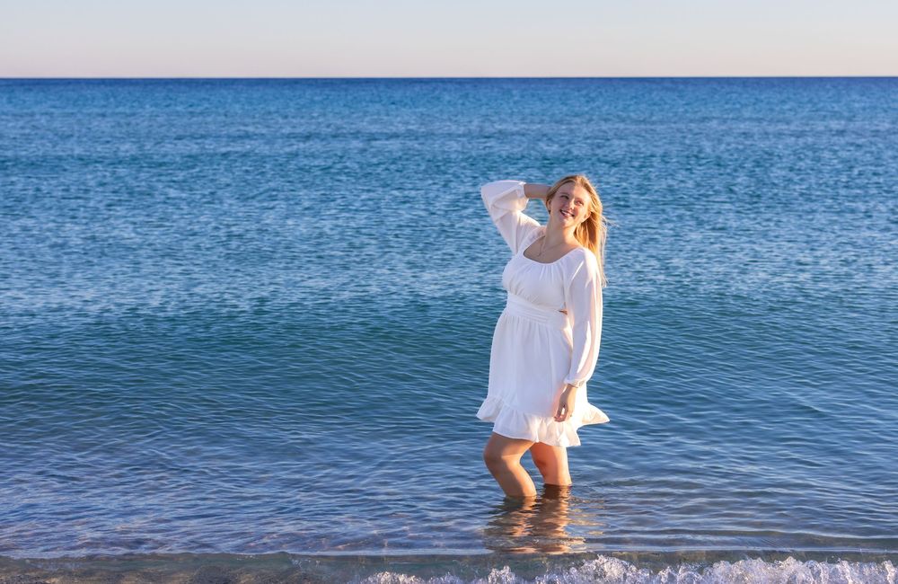 A person in a flowing white dress stands ankle-deep in blue ocean waters on a sunny day.
