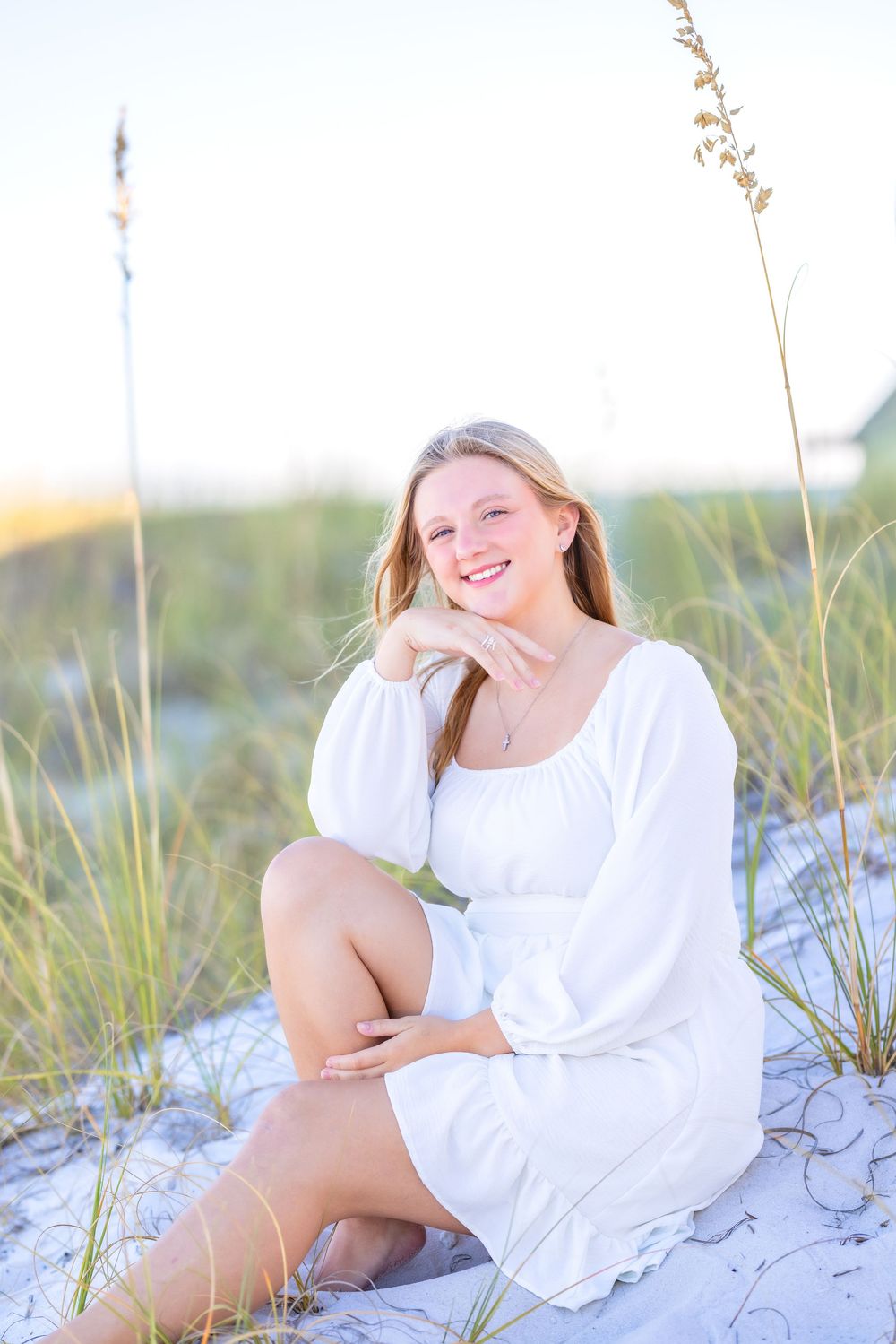 Person in a white dress poses on sandy dunes with sea grass at sunset creating a dreamy beach portrait.