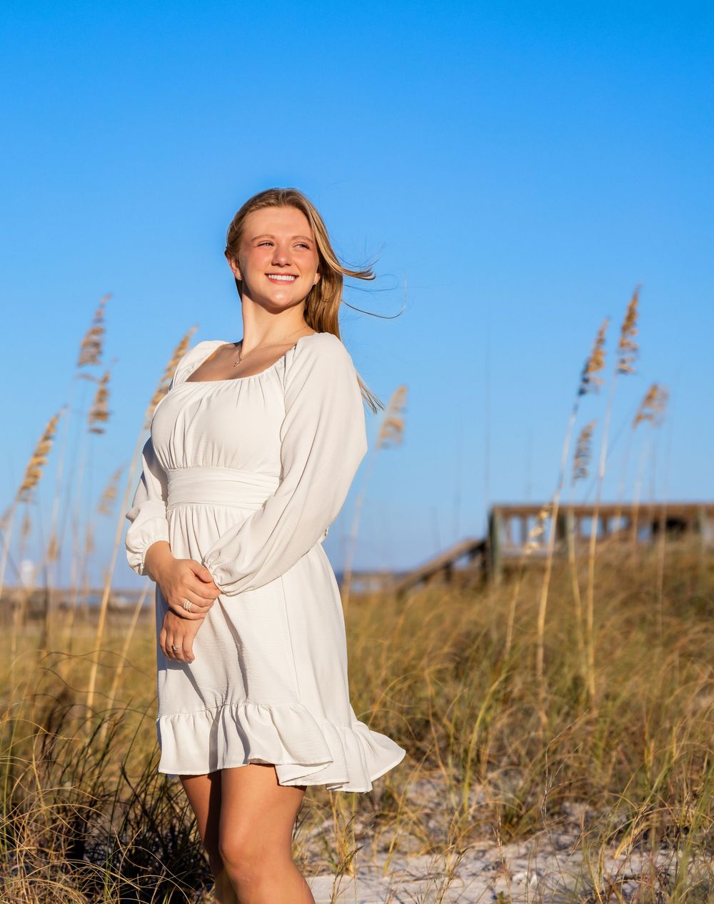A person in a white flowy dress poses against tall beach grass and blue sky on a sunny day.