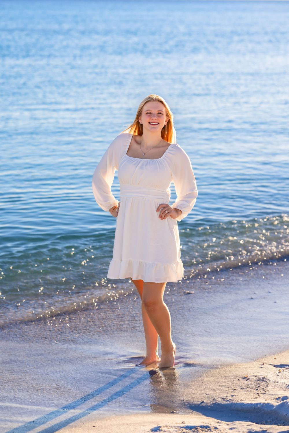 Person in white dress plays and dances along the shoreline as waves gently lap at the beach.