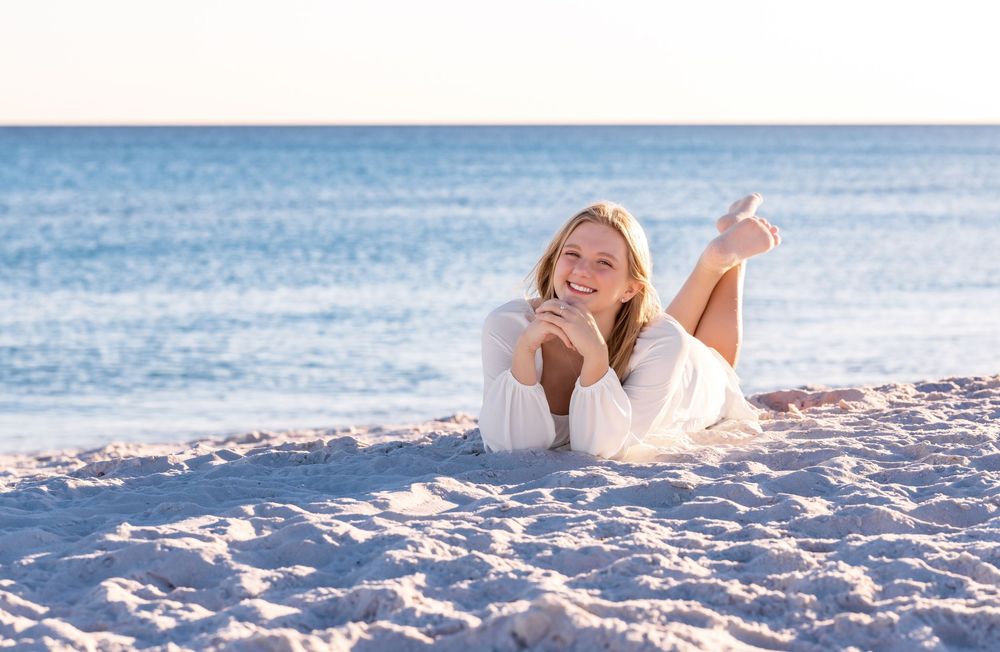 A person in a white sweater relaxes on a sandy beach near blue ocean waters during a sunny day.