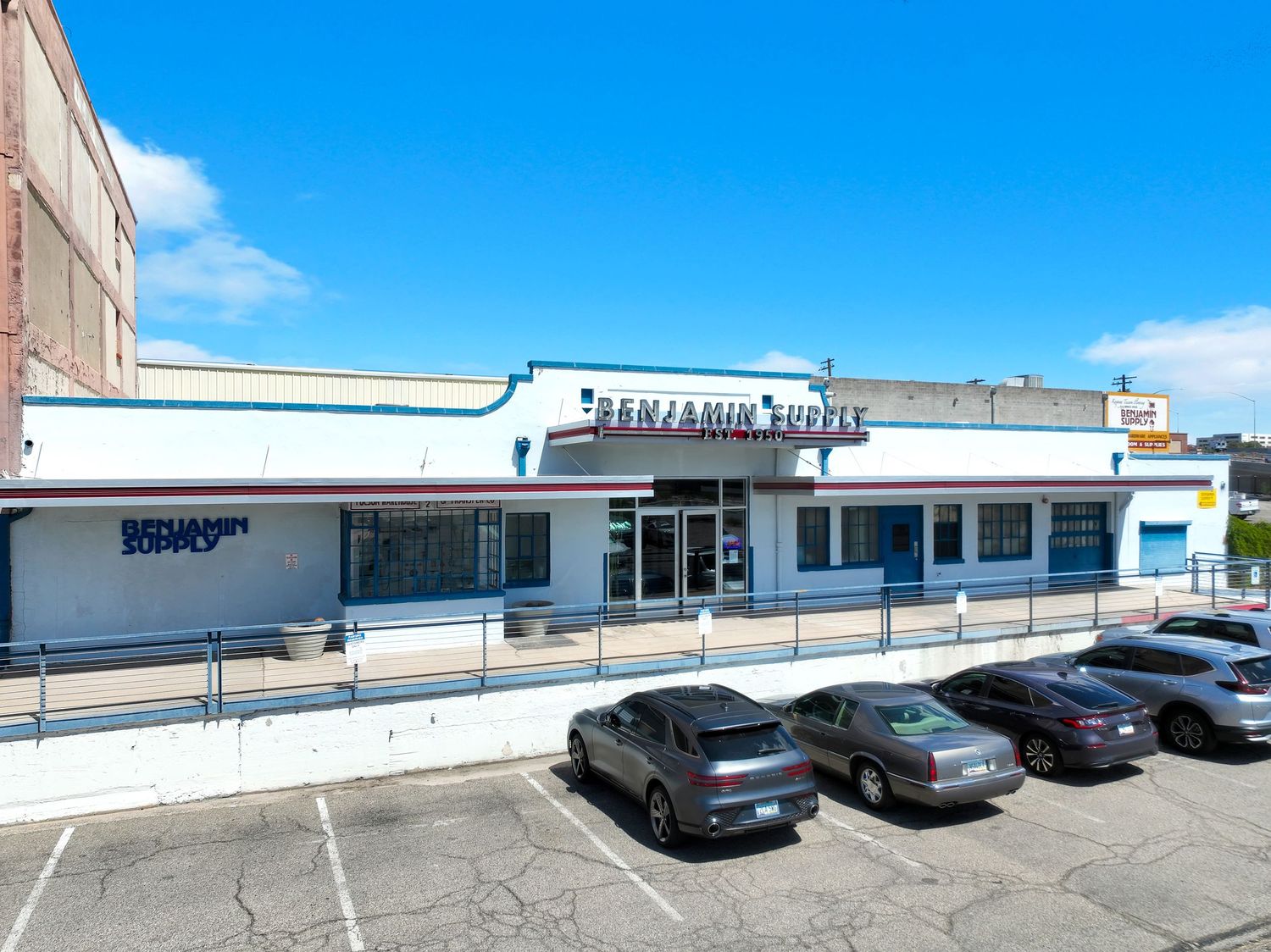 Long white commercial strip mall building with parking lot under bright blue sky.