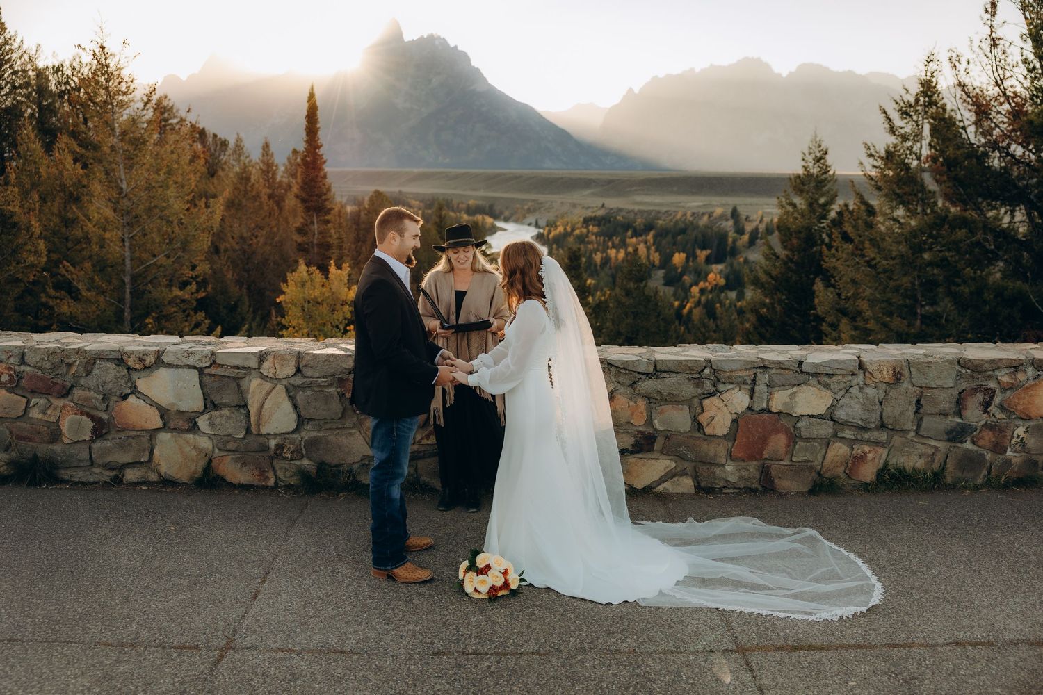 A couple shares a romantic moment at sunset against a mountain backdrop with a stone wall and autumn foliage.