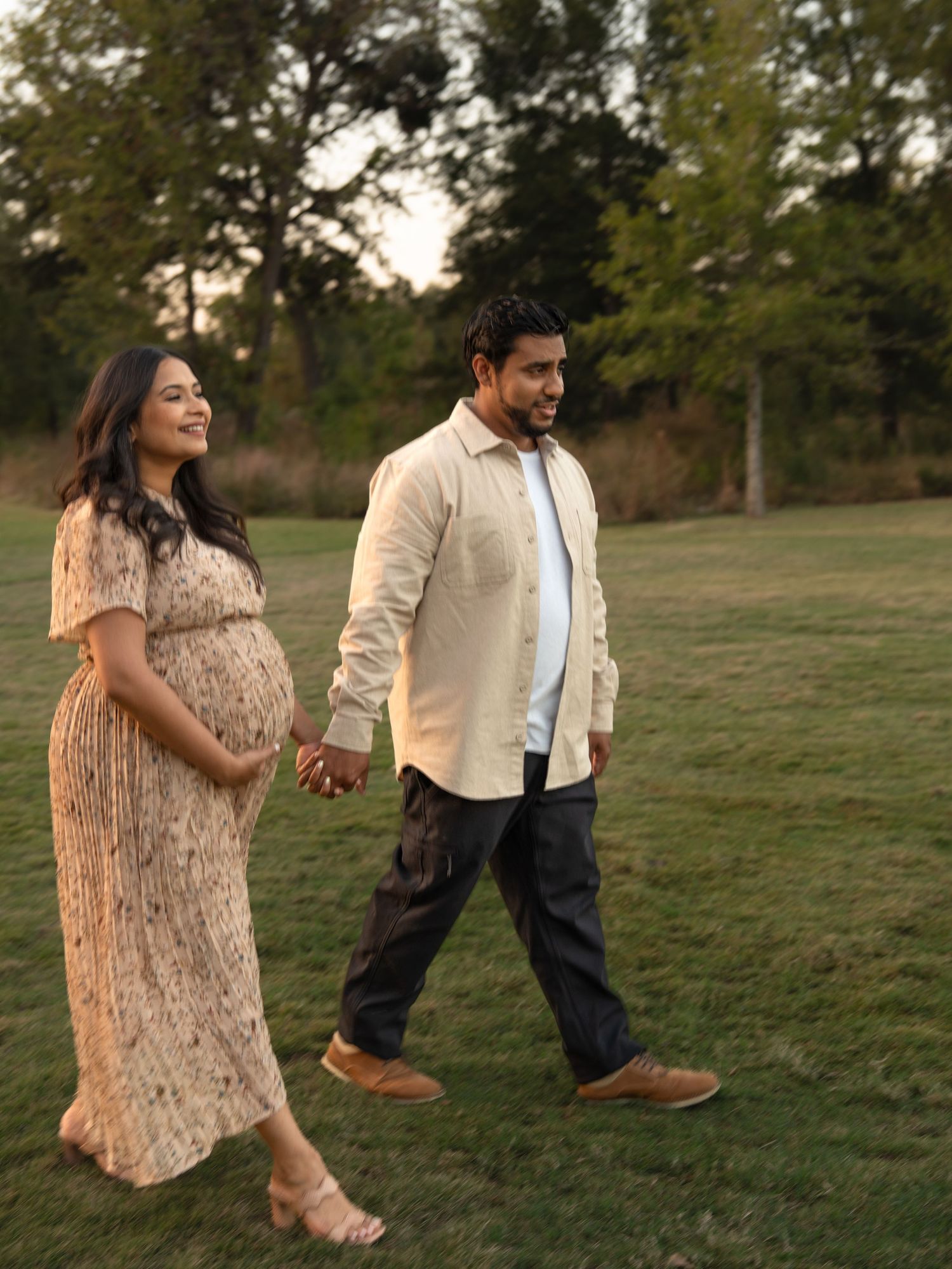 A couple takes a romantic stroll through a grassy field at sunset, with the woman wearing a flowing beige dress.
