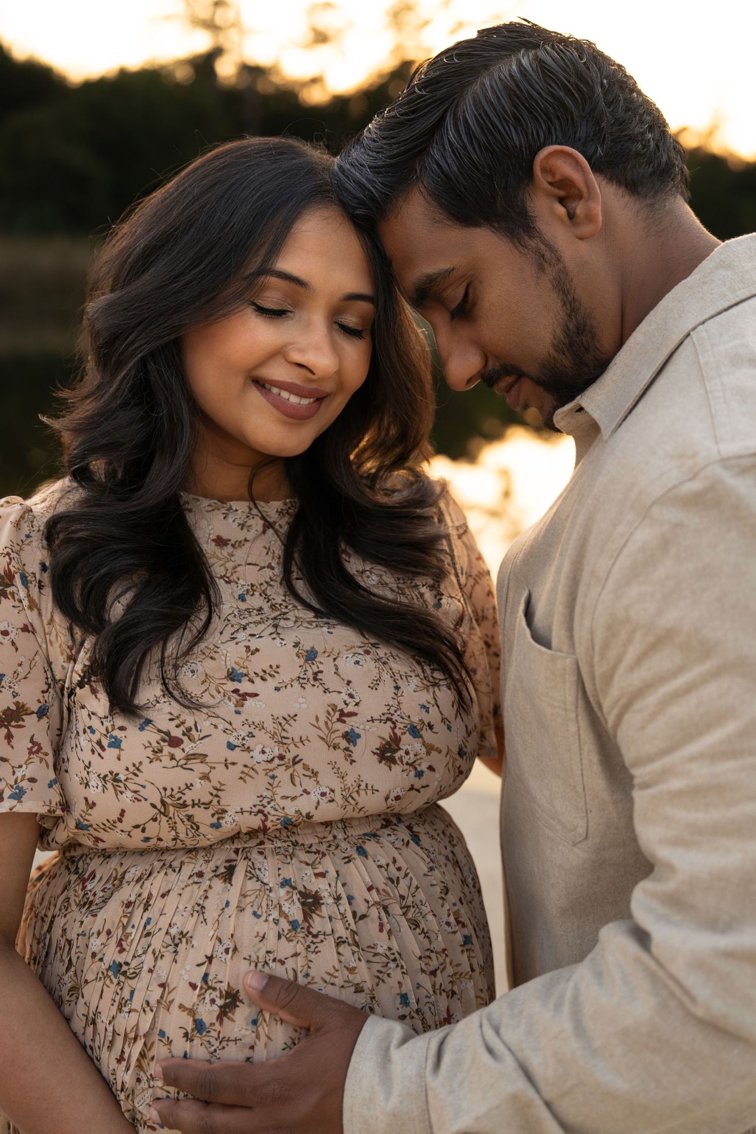 Intimate maternity photoshoot at sunset shows couple in floral dress and cream shirt embracing tenderly.