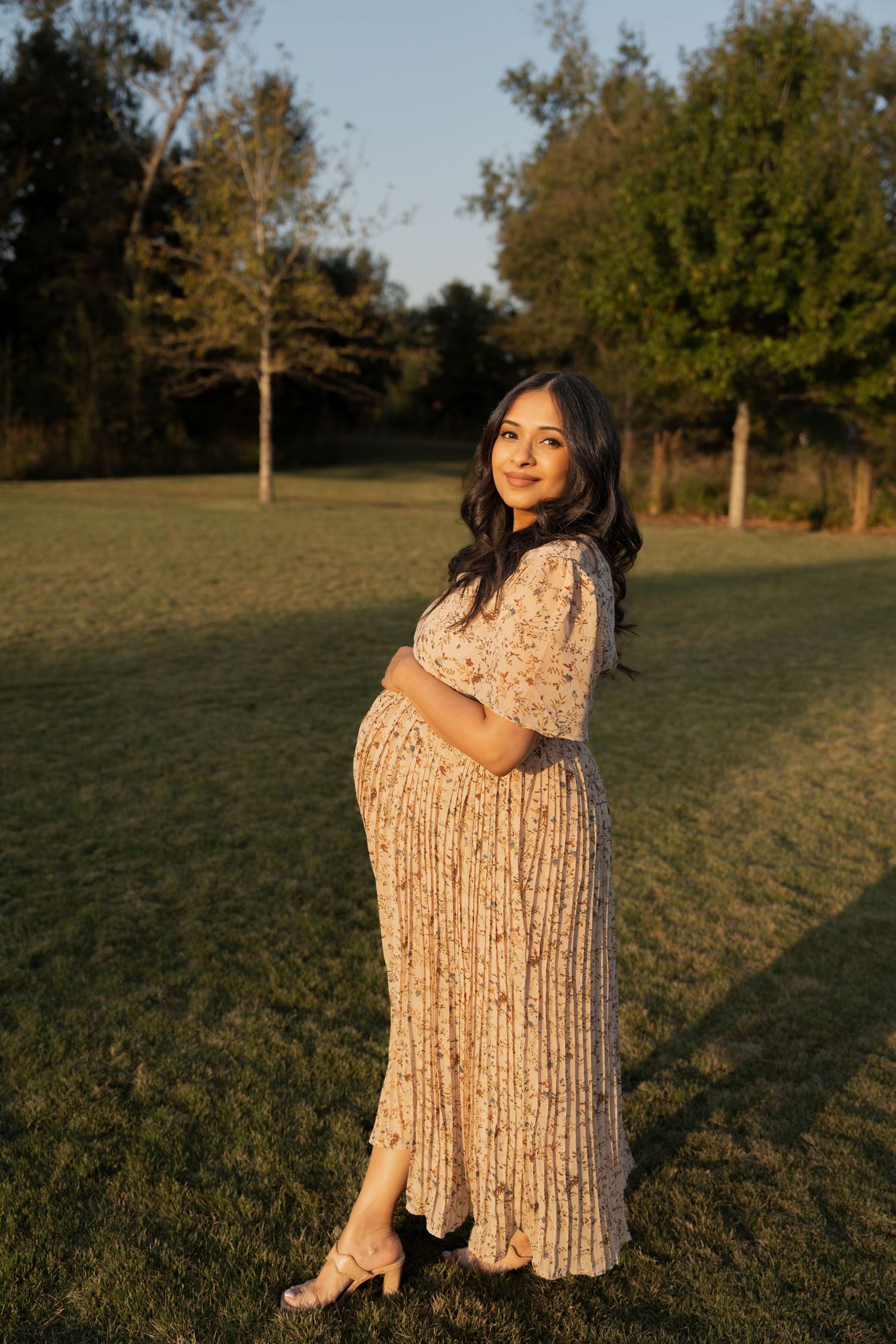 Woman in a floral dress poses gracefully on grass during golden hour outdoor photography session.