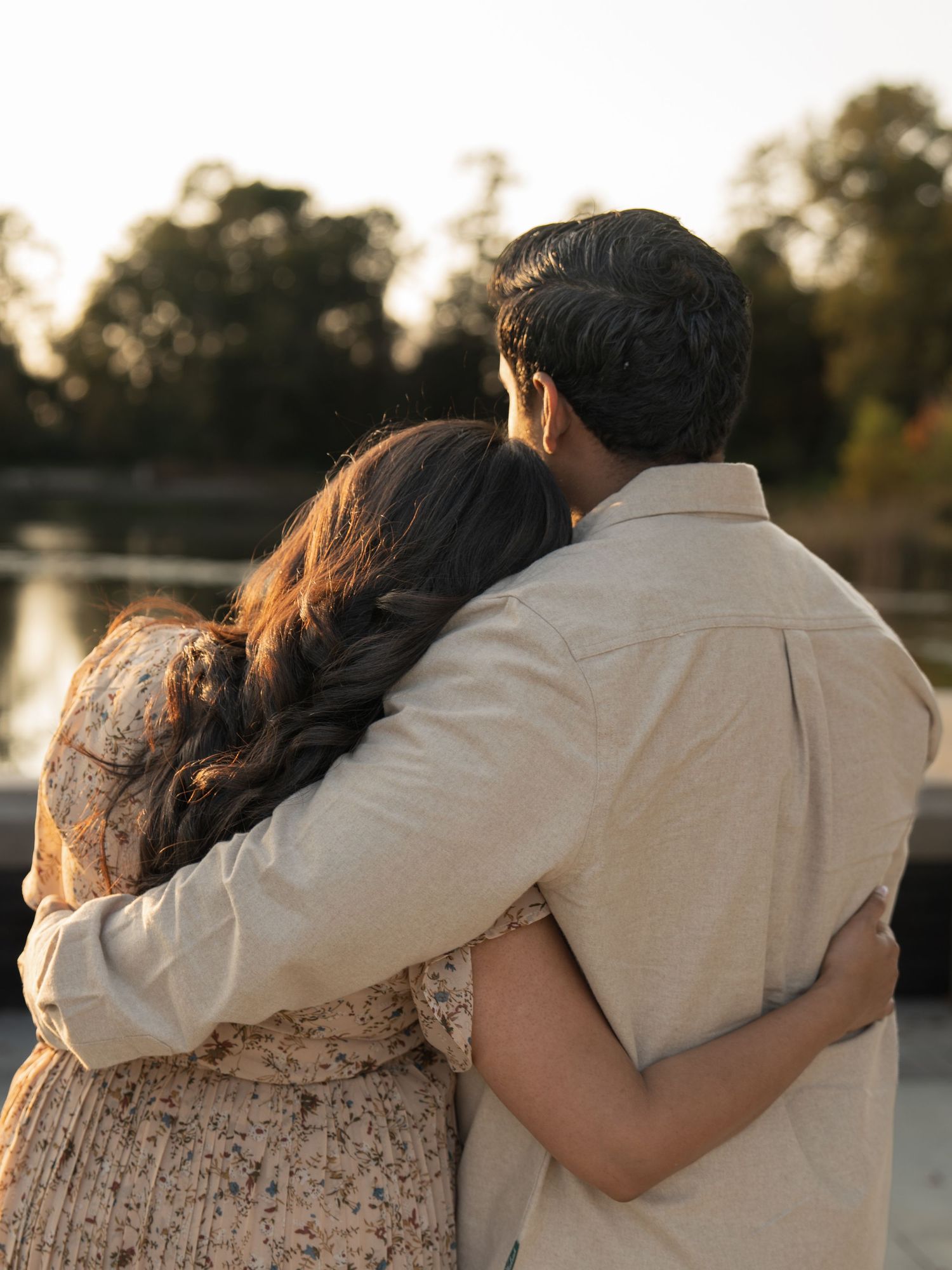 A couple embraces while watching the sunset by a lake, with the person in white holding their partner close.