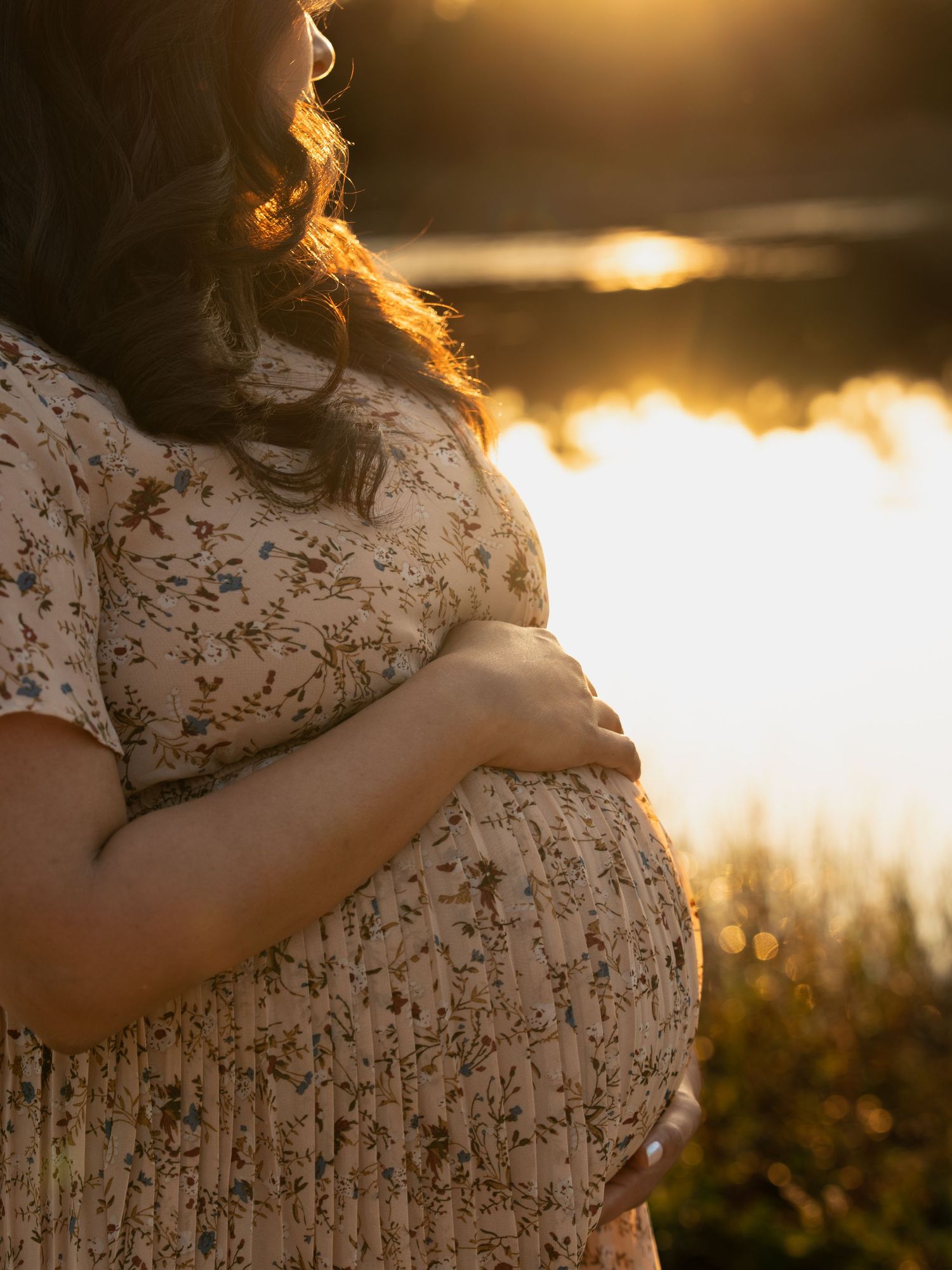 A pregnant woman in a floral dress holds her belly while standing in golden sunset light by a lake.