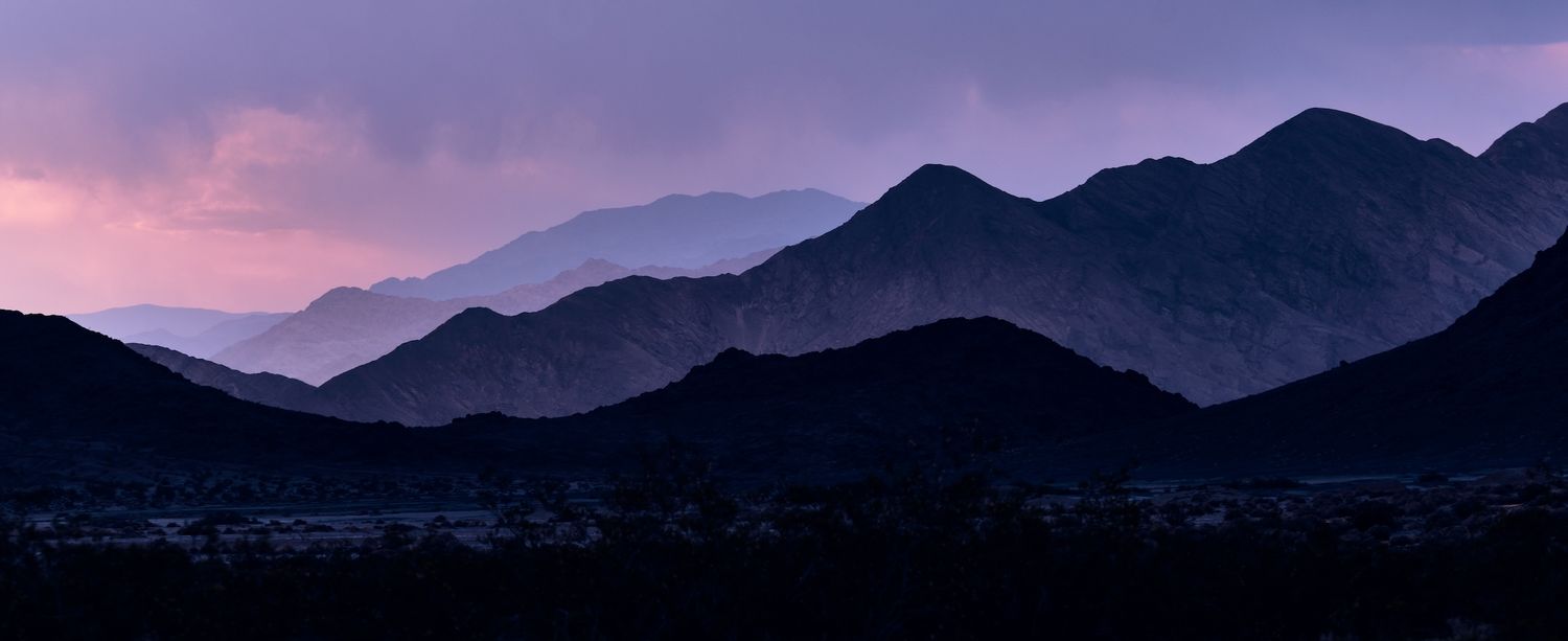 Purple twilight panorama of layered mountain silhouettes with small town lights at base of range.