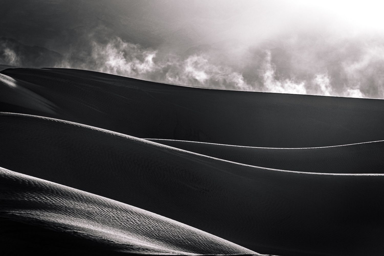 Black-and-white desert sand dunes, backlit by a dramatic dust stream, creating a striking contrast of light and shadow.