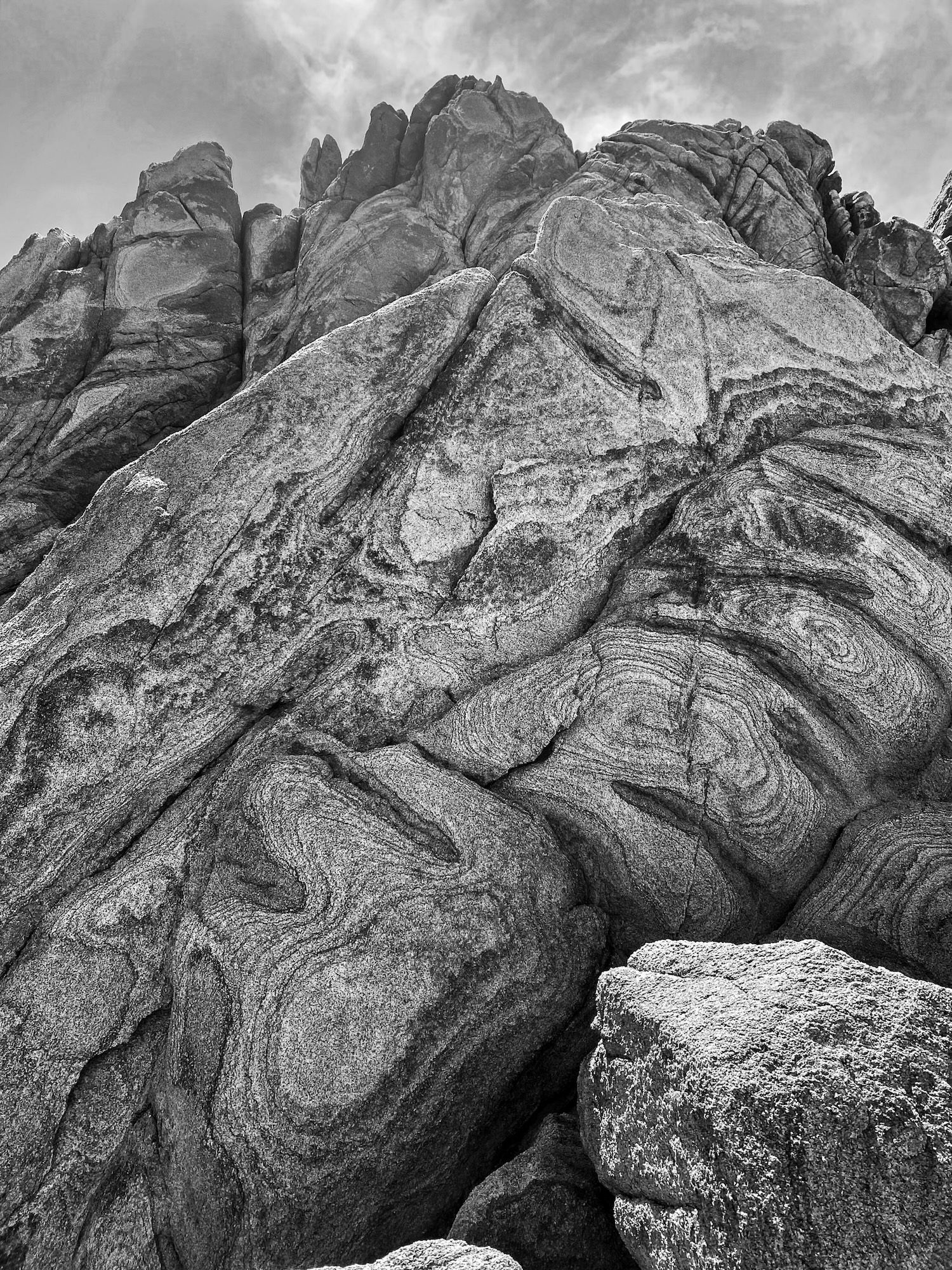 Black-and-white photo of swirling granite rock formations with dramatic layered patterns against a cloudy sky.