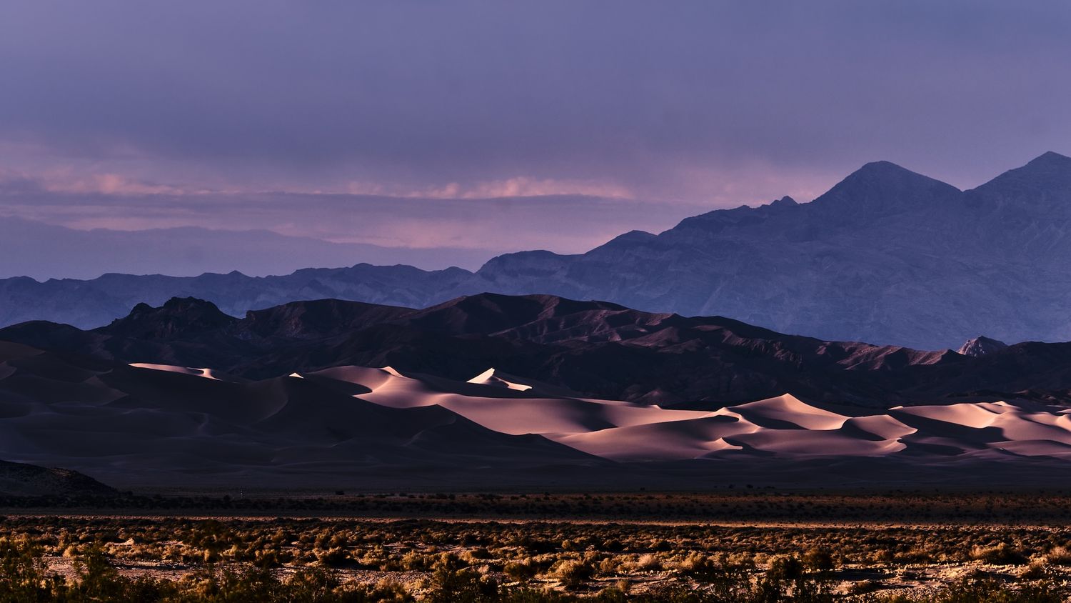 Glowing sand dunes at twilight with dramatic purple storm clouds over distant mountain range in Death Valley.