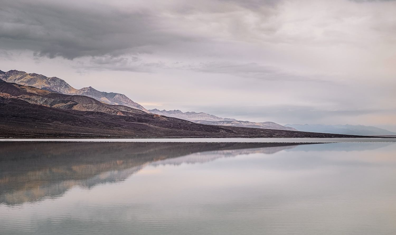 Serene still water reflecting overcast sky and rugged mountain range in Death Valley National Park.