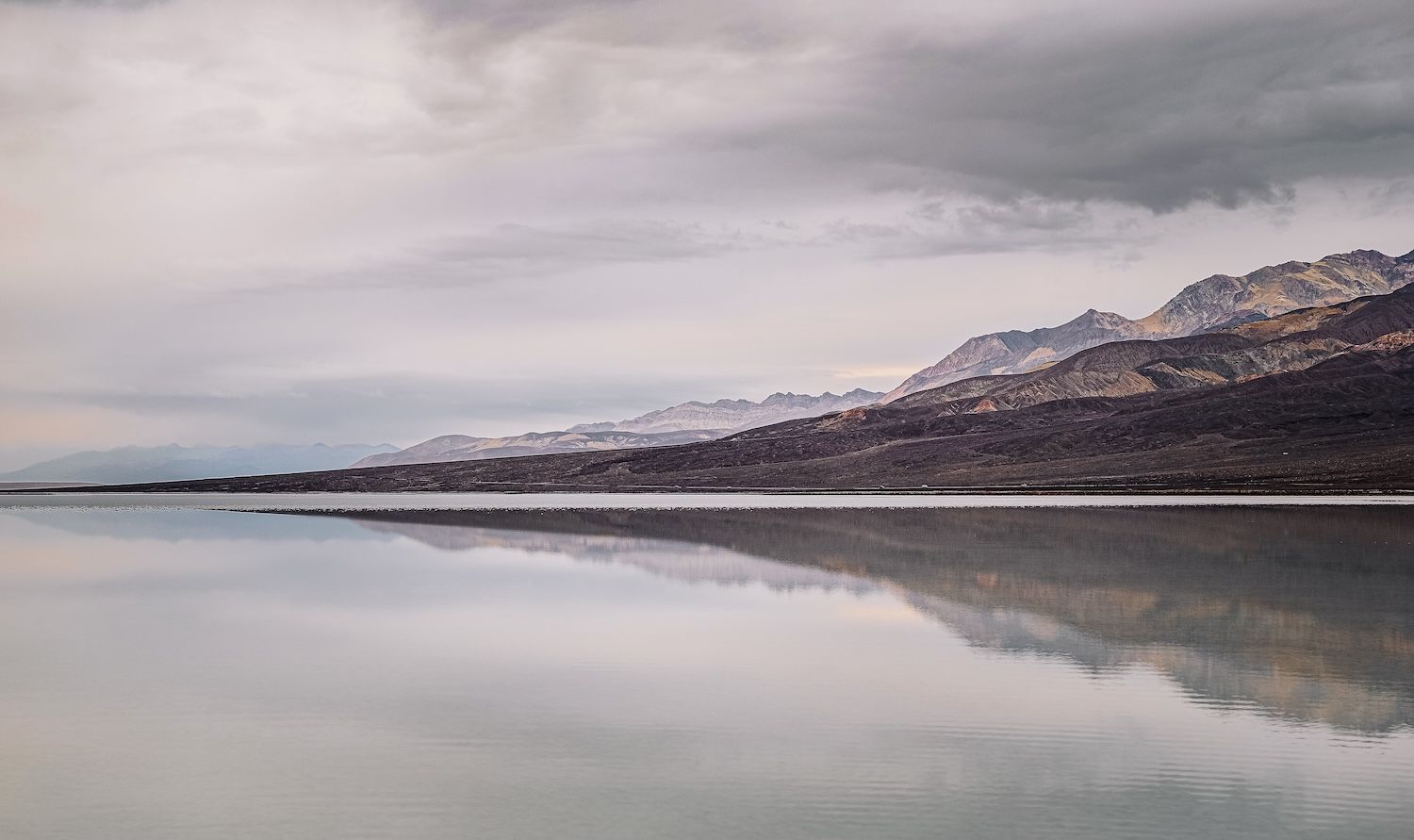 Calm reflective lake mirroring cloudy sky and distant mountains in Death Valley desert landscape.