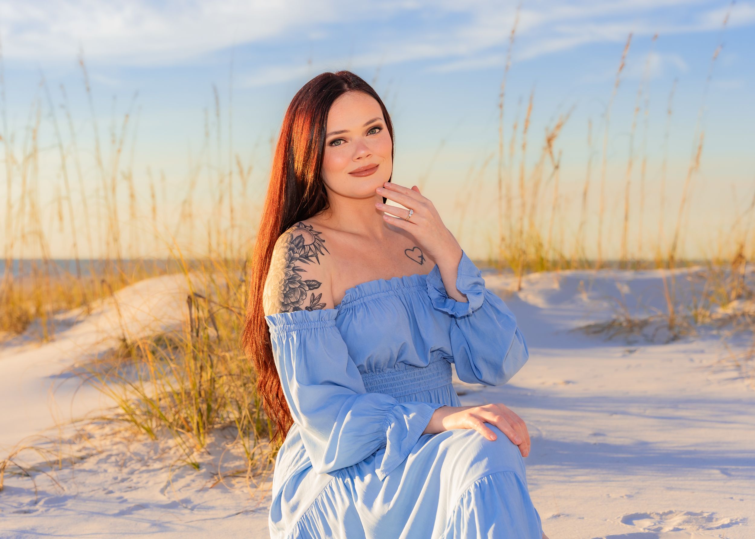 A woman in a blue off-shoulder dress sits on white sand dunes at sunset, with sea oats in the background.