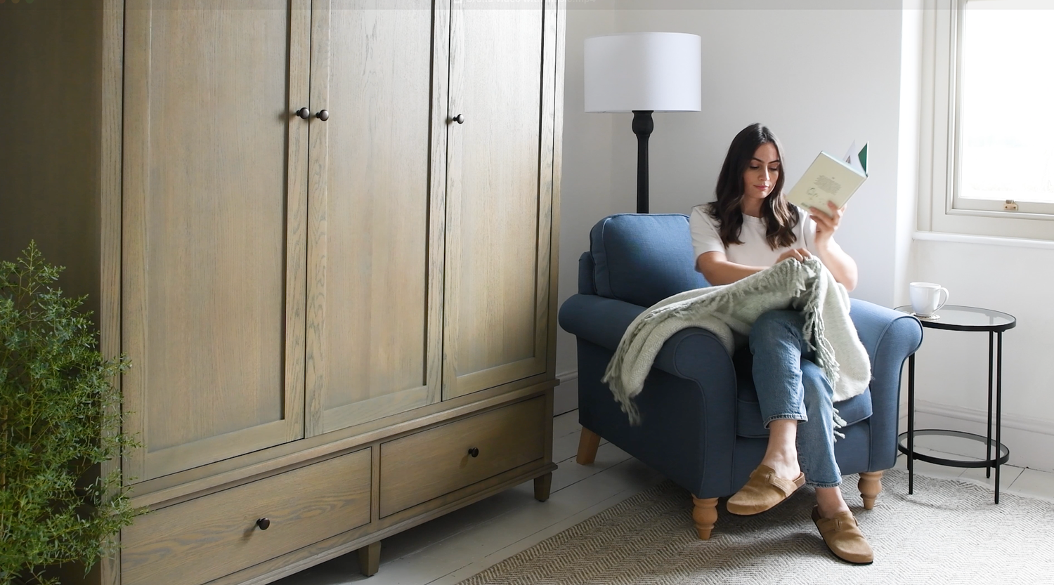 Someone sits comfortably in a blue armchair reading a book next to a wardrobe in a cozy home setting.