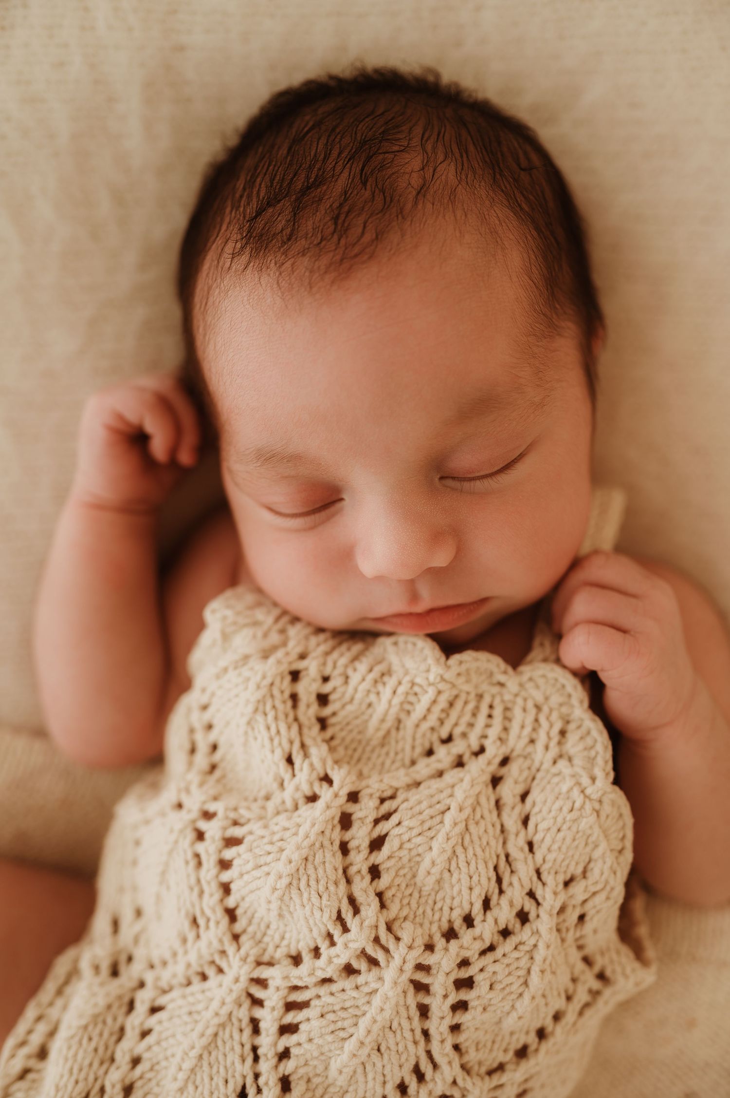 Newborn baby peacefully sleeping while wrapped in soft white knitted blanket on cream colored background.