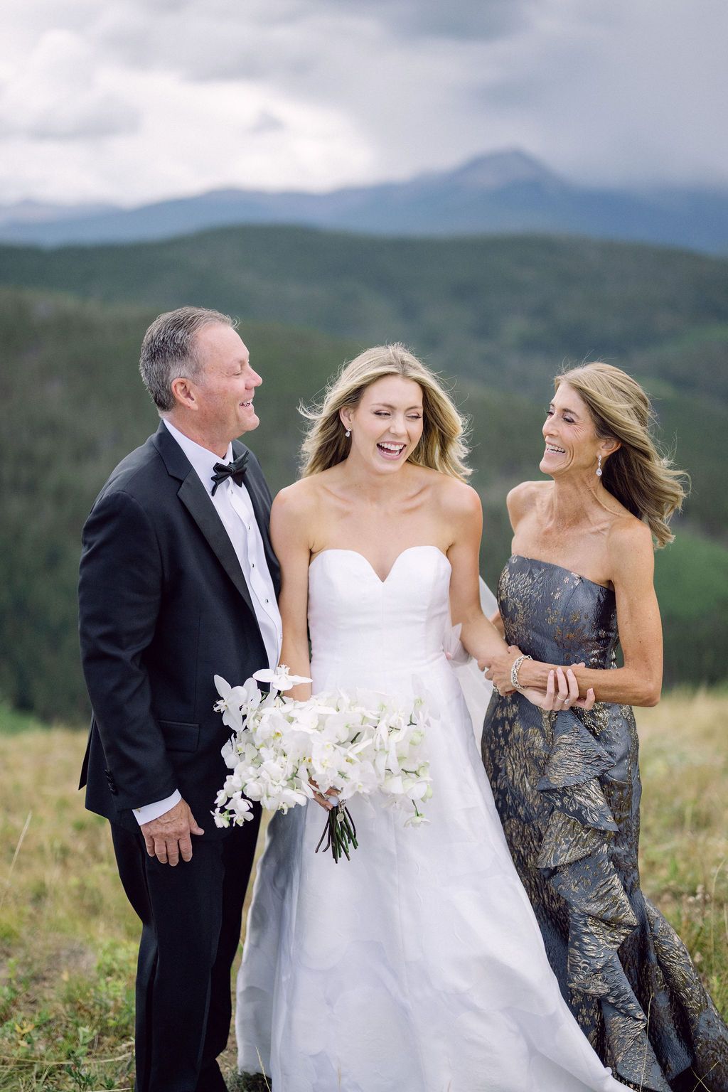 Two people embrace and smile while posing outdoors with mountains in the background and white flowers visible.