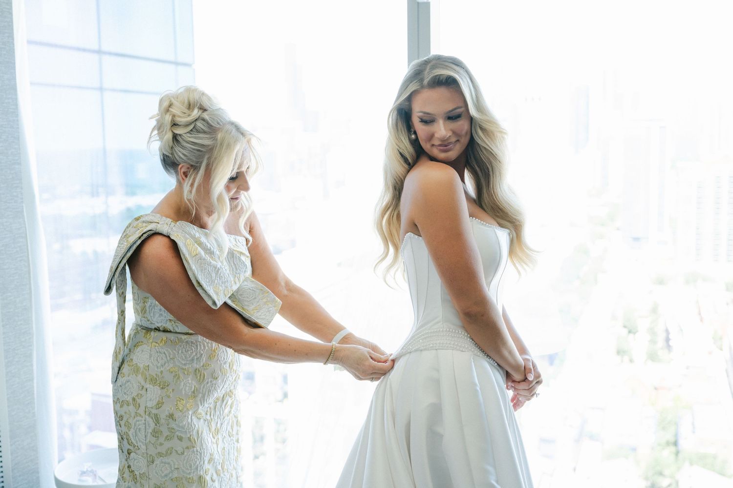 A bride and bridesmaids help with final dress adjustments in a bright sunlit room before the wedding ceremony.