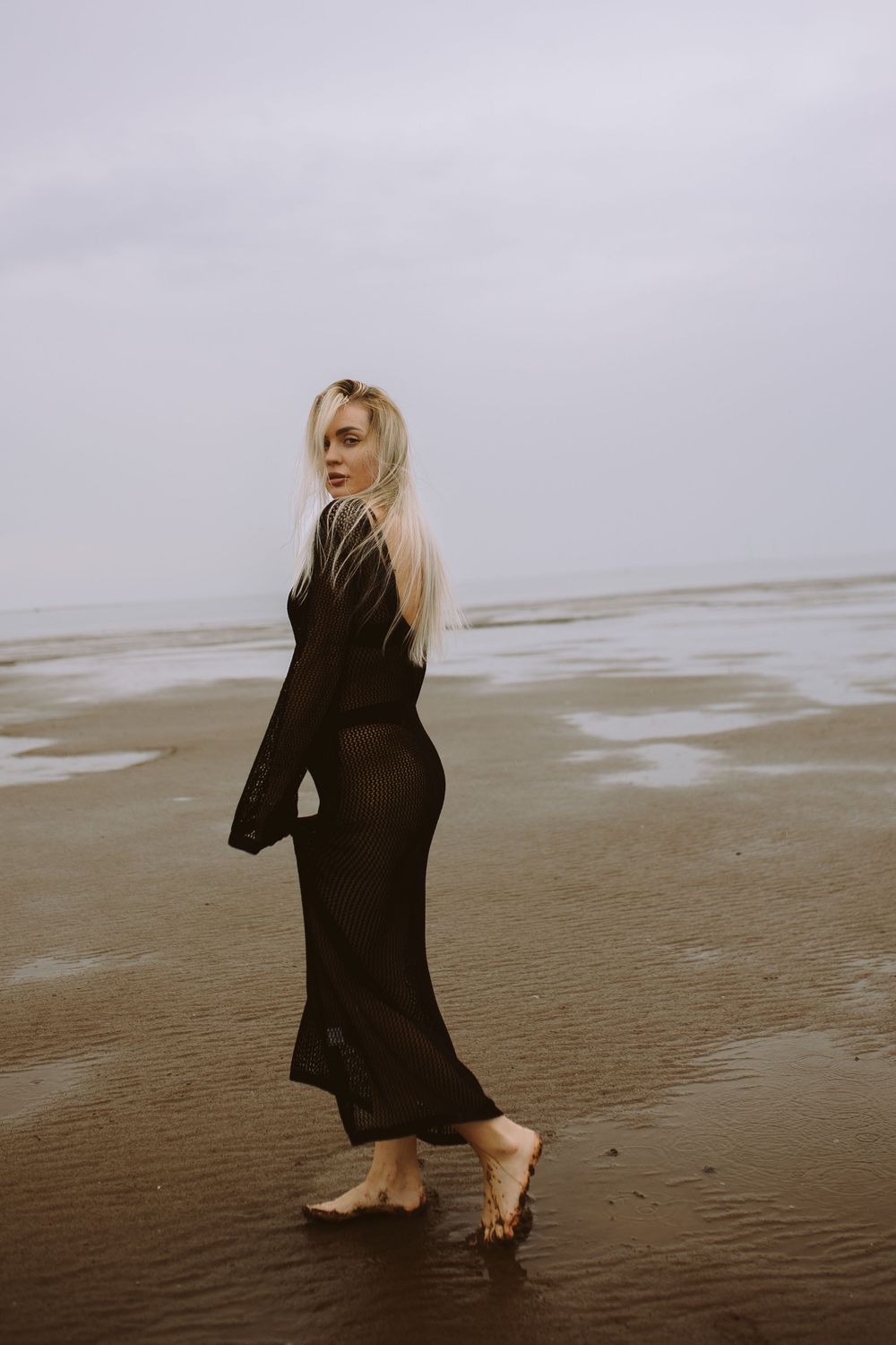A person in a black dress poses on a misty beach at low tide during an overcast day.
