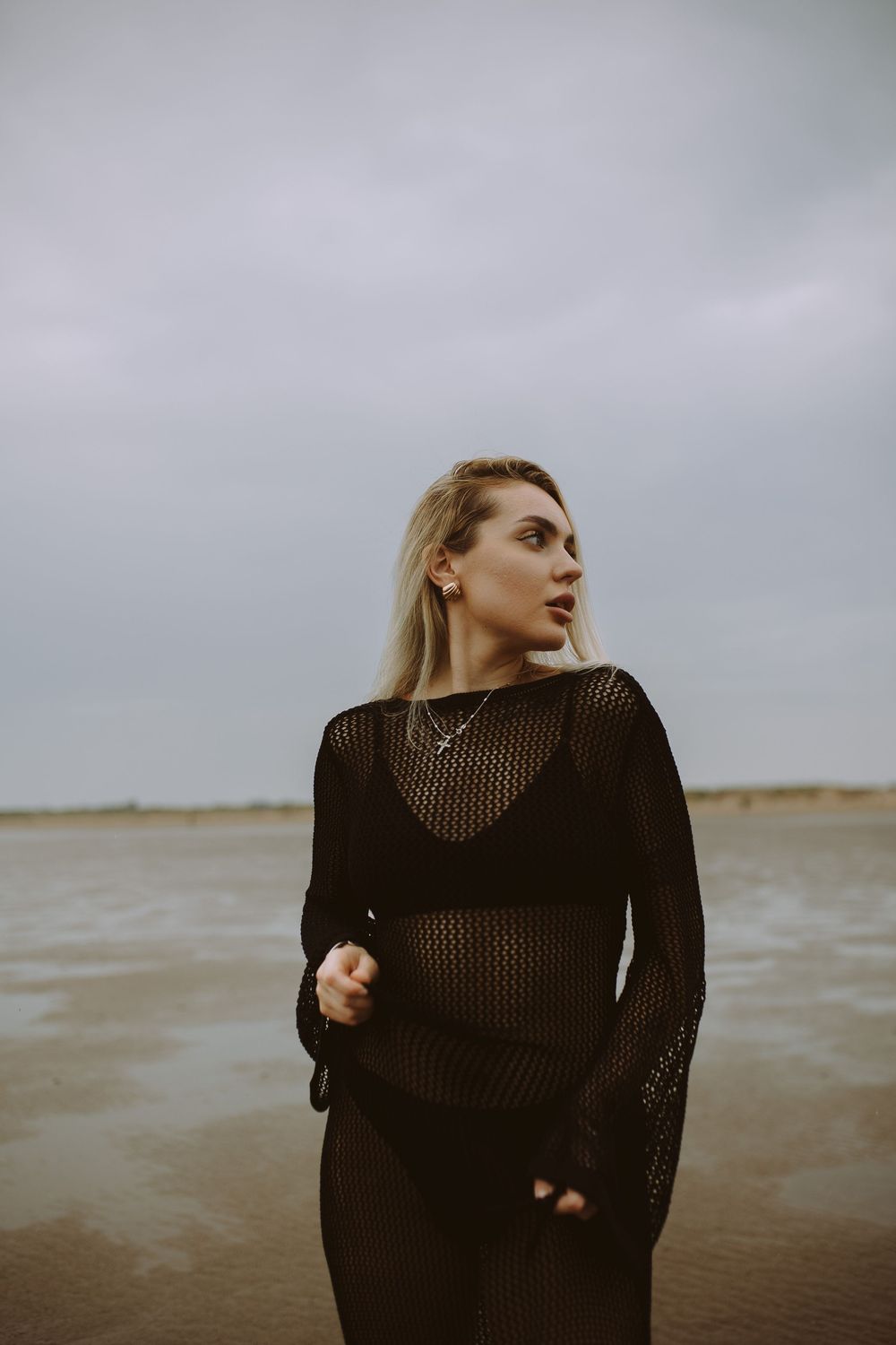 Moody series of photos showing a person in a black mesh sweater standing against a gray ocean backdrop.