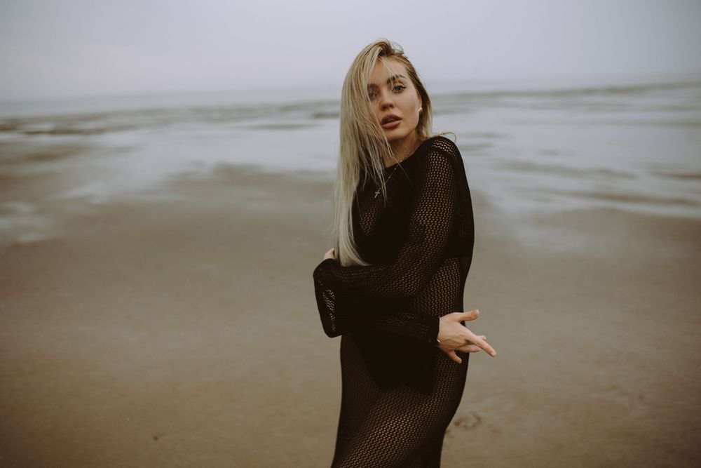 A person in a dark dress walks along a misty beach shoreline at dusk creating a moody atmospheric scene.