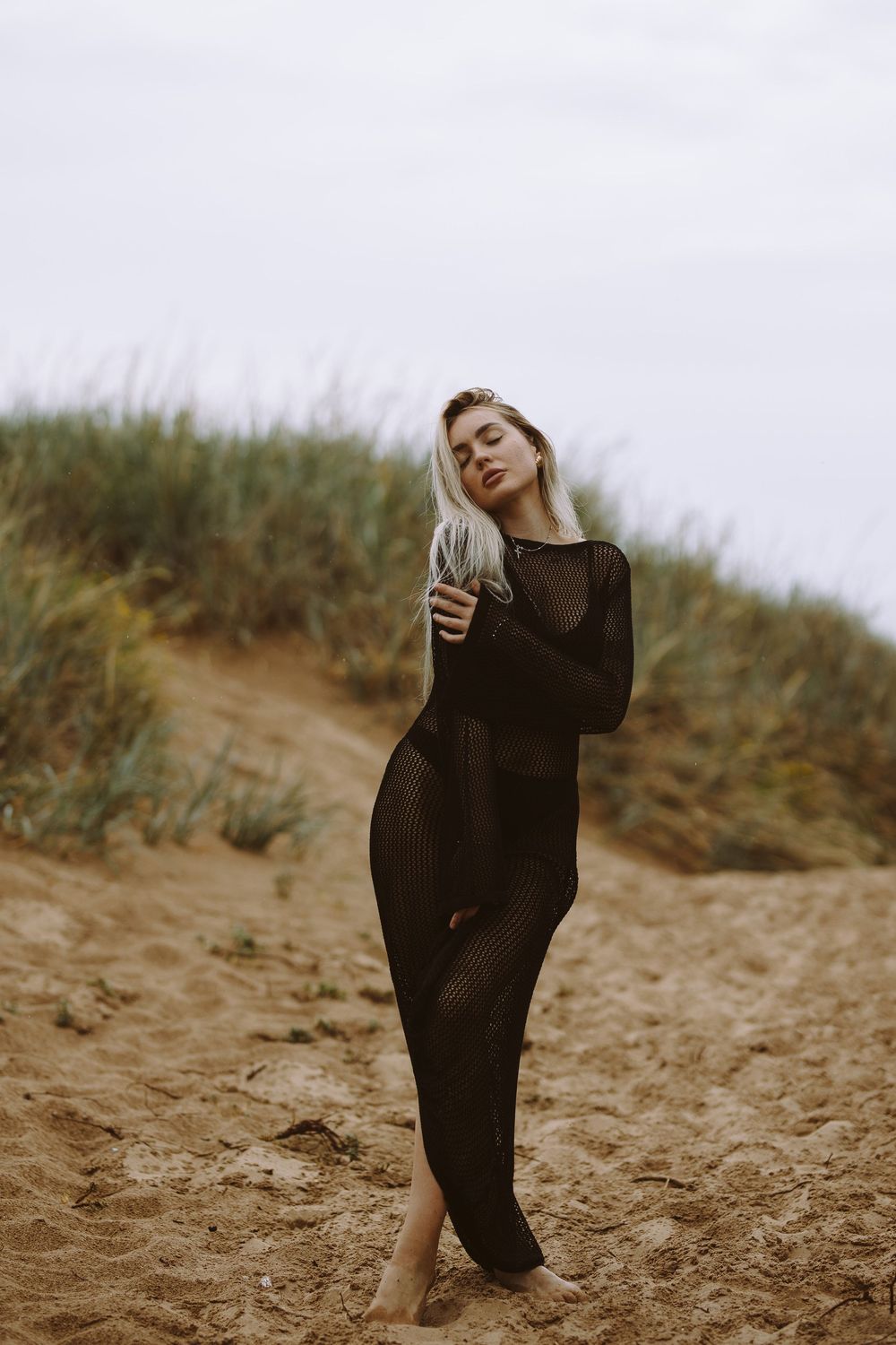 A person in a flowing black dress poses gracefully on a sandy beach dune during a moody outdoor photoshoot.