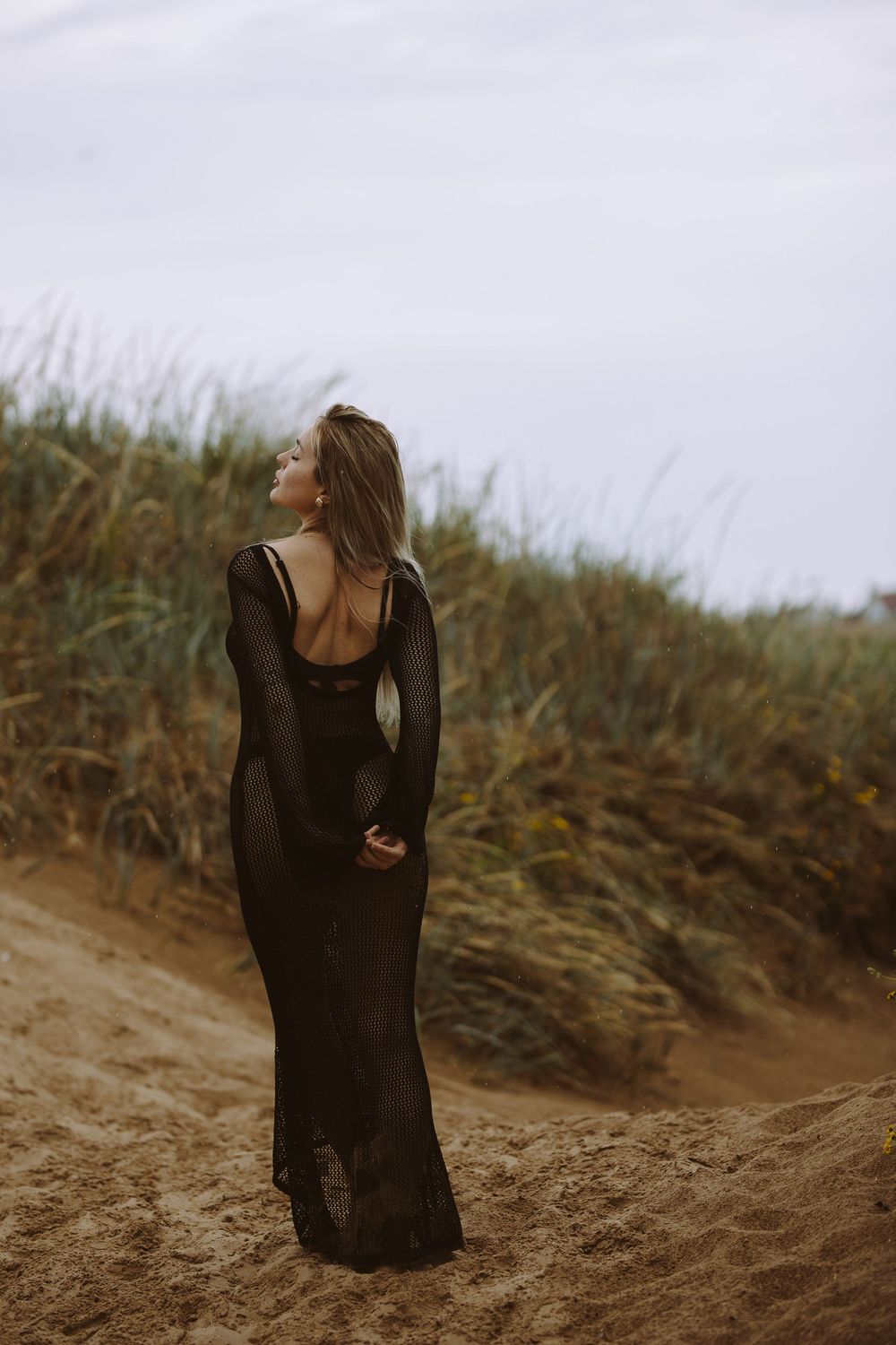 A person in a flowing black dress poses dramatically on sandy dunes against a moody cloudy sky.