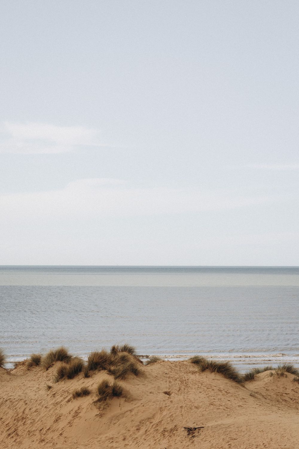Scenic coastal landscape with sandy dunes, beach grass, and calm ocean waters under a bright cloudy sky.