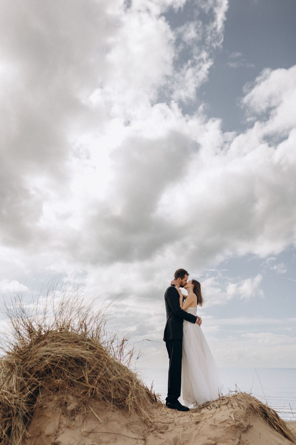 A romantic wedding photo series showing a couple embracing on a sandy dune against a cloudy sky.