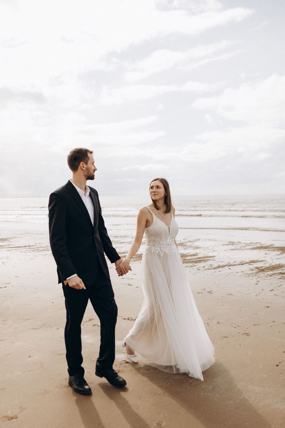 A couple in wedding attire walking hand in hand on a cloudy beach with waves in the background.