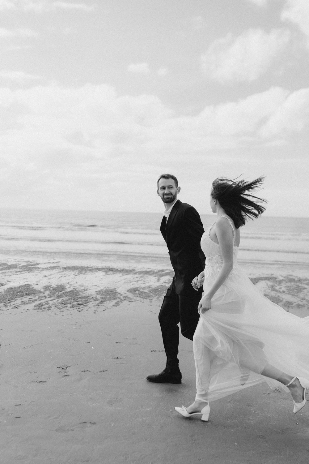 A couple shares a playful moment running and dancing together on a windy beach in black and white photography.